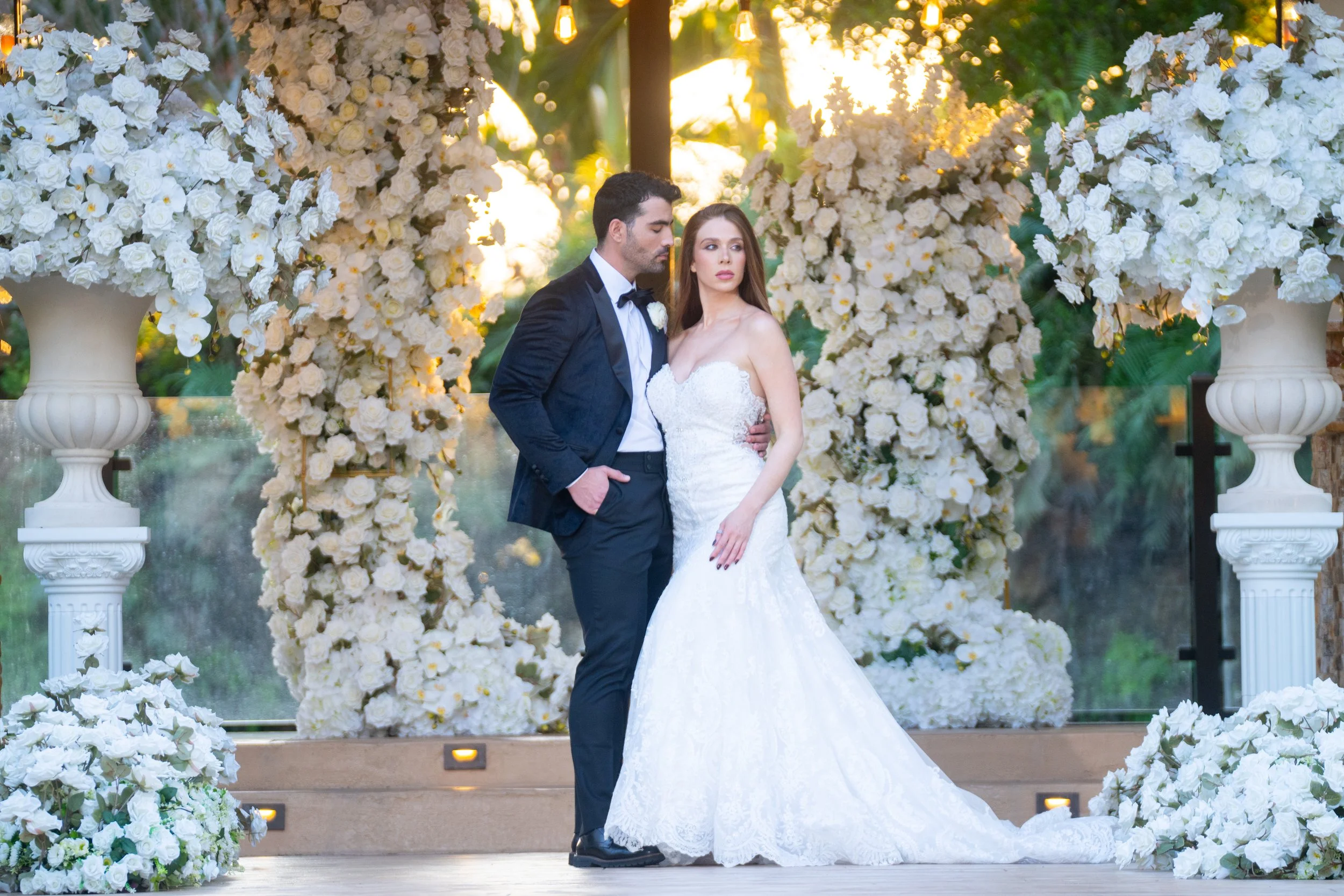 A bride and groom standing close together at a wedding ceremony, surrounded by large white floral arrangements and greenery, with warm sunlight in the background.