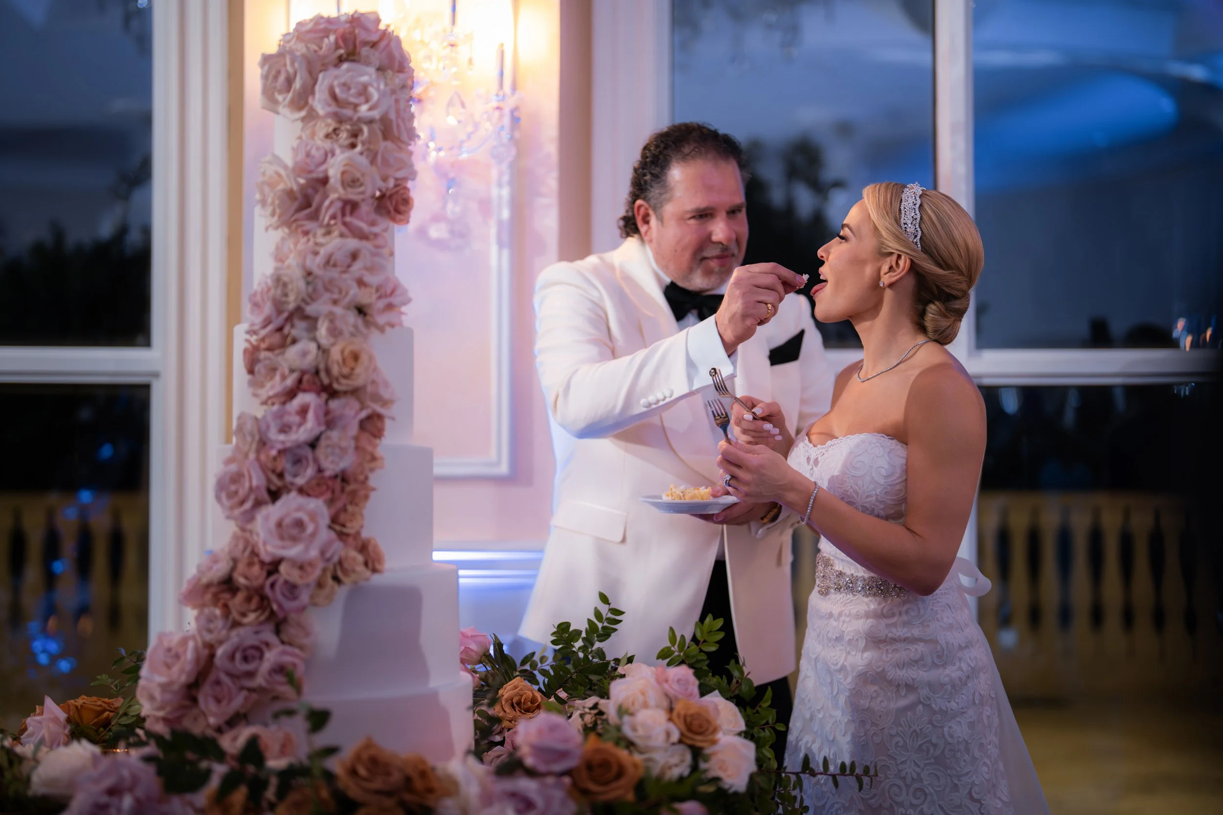 A bride and groom at their wedding reception, with a groom feeding a piece of cake to the bride. There is a large wedding cake decorated with pink and peach roses and greenery, and a floral arrangement underneath it. They are standing inside near lar