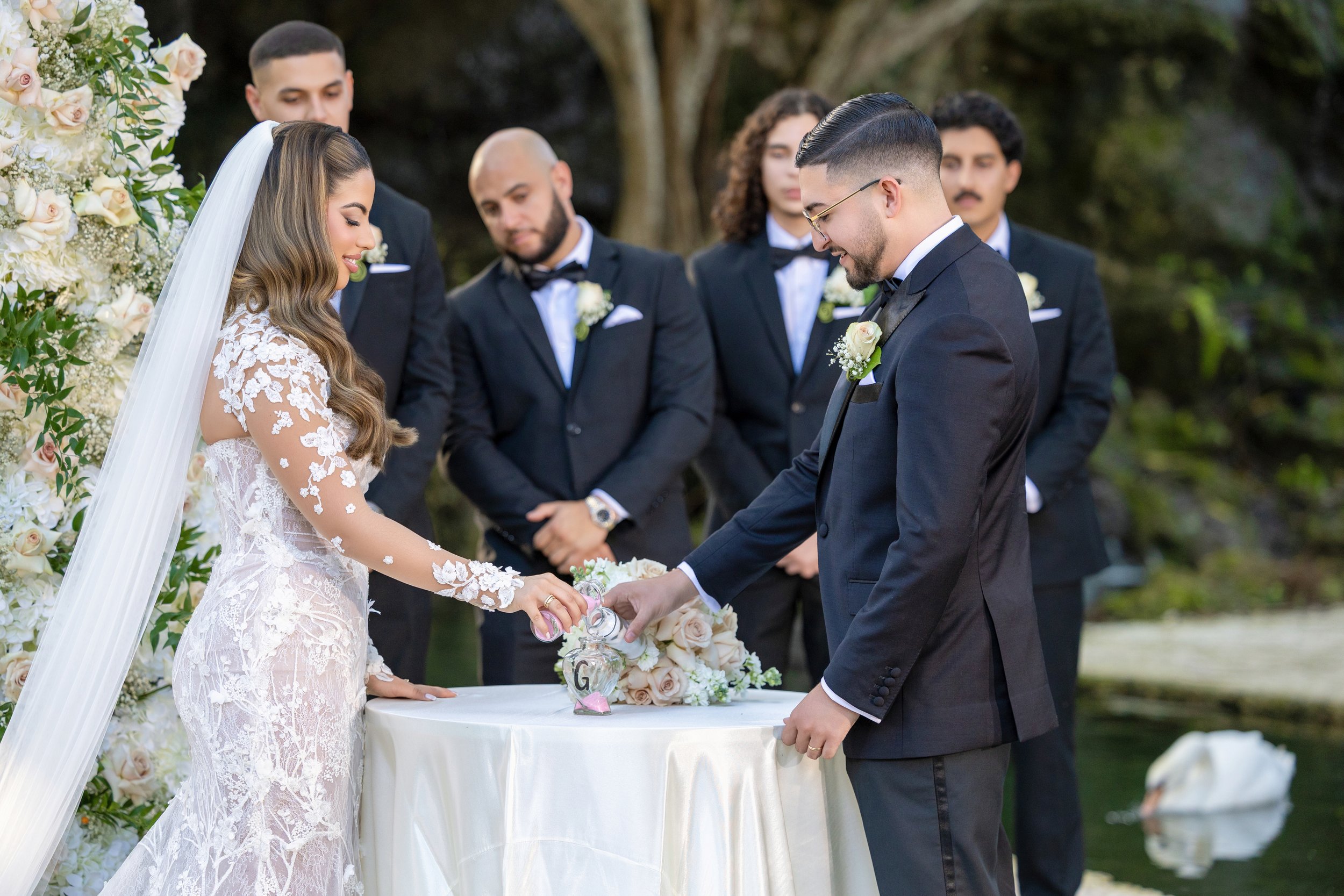 Bride and groom exchanging wedding rings under an outdoor floral arch, with groomsmen standing behind them.
