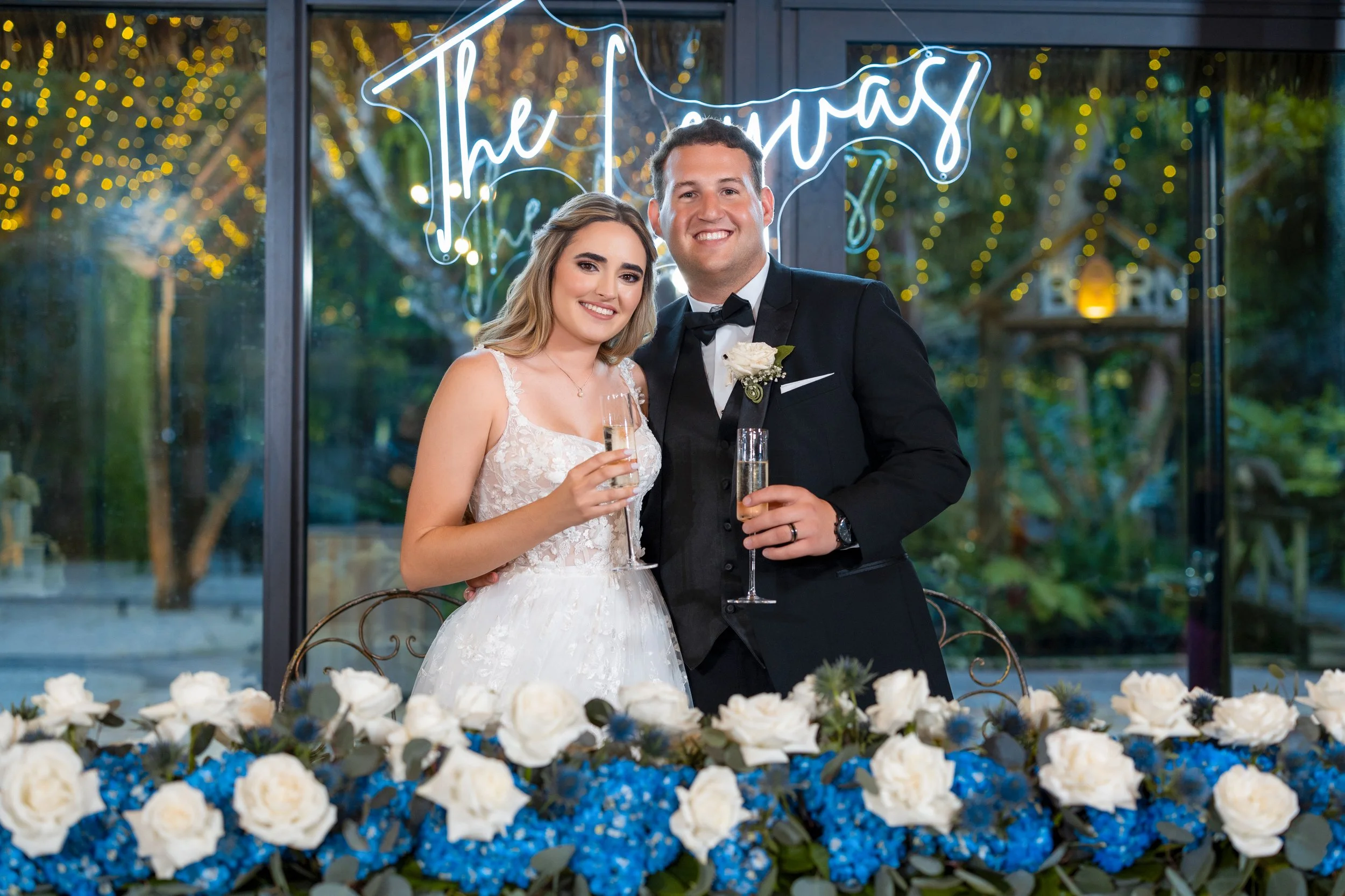 Bride and groom at wedding reception, standing by a decorated table with white roses and blue flowers, holding champagne glasses and smiling inside a venue with a neon sign that reads 'The Memories' and string lights in the background.