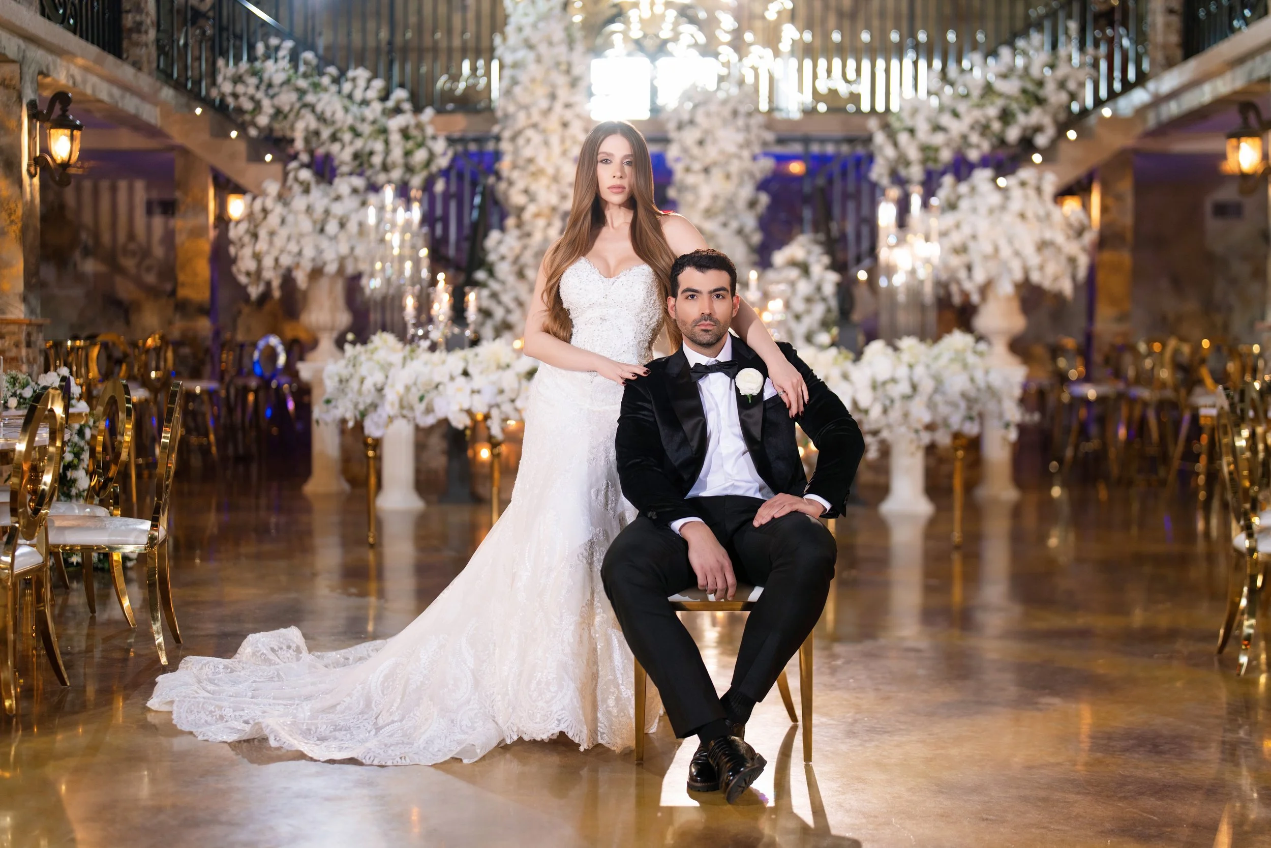 A bride and groom in wedding attire at a decorated wedding venue with white flowers and gold chairs.