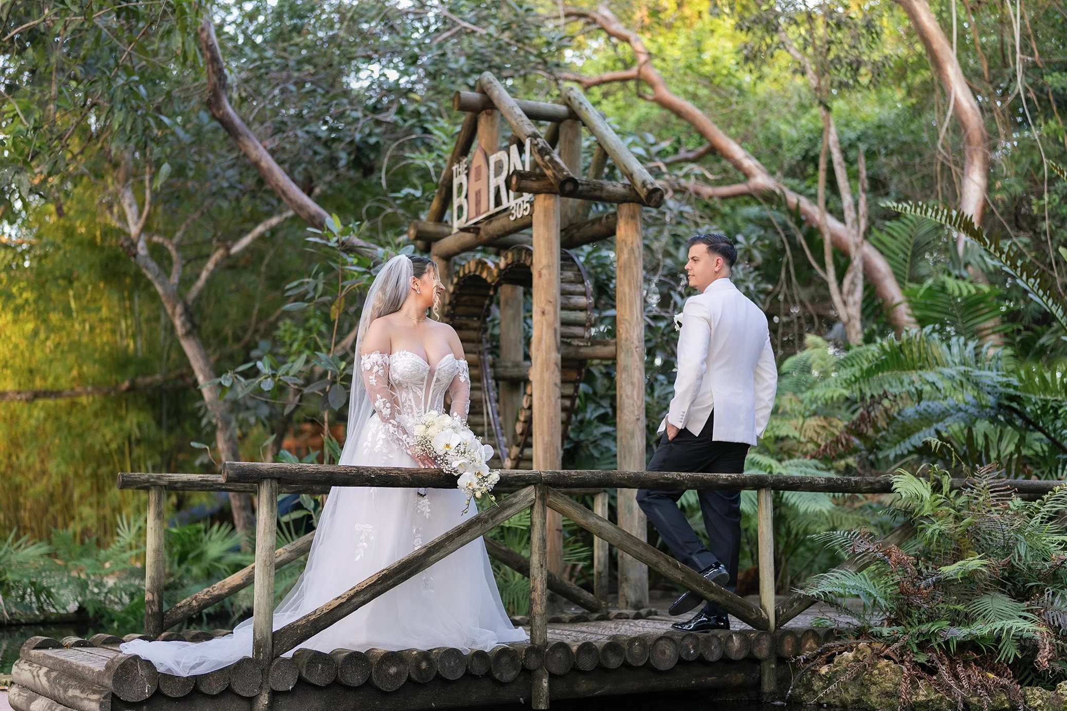 A bride and groom standing on a wooden bridge in a lush, green outdoor setting with trees and plants, exchanging looks during their wedding ceremony.