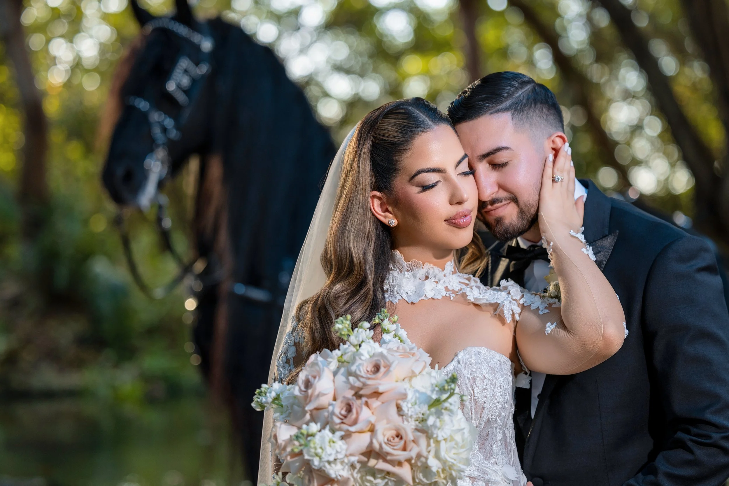 A bride and groom share an intimate moment outdoors with a black horse in the background. The bride is holding a bouquet of pink and white roses and is wearing a lace wedding dress, while the groom is dressed in a black tuxedo. They are embracing ten