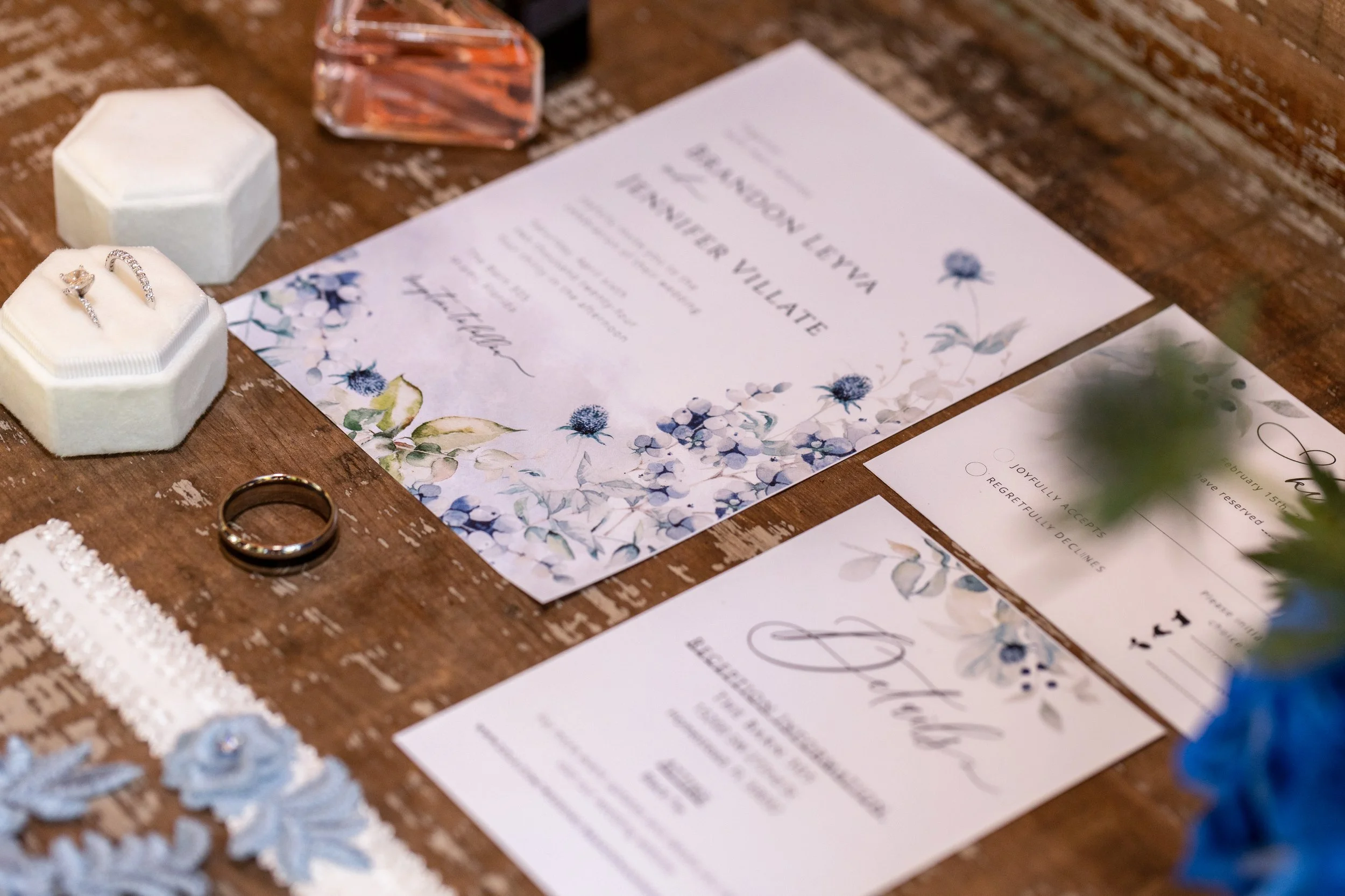 Wedding invitation suite, wedding rings, lace garter, and jewelry on a wooden table, with a blue floral bouquet partially visible.