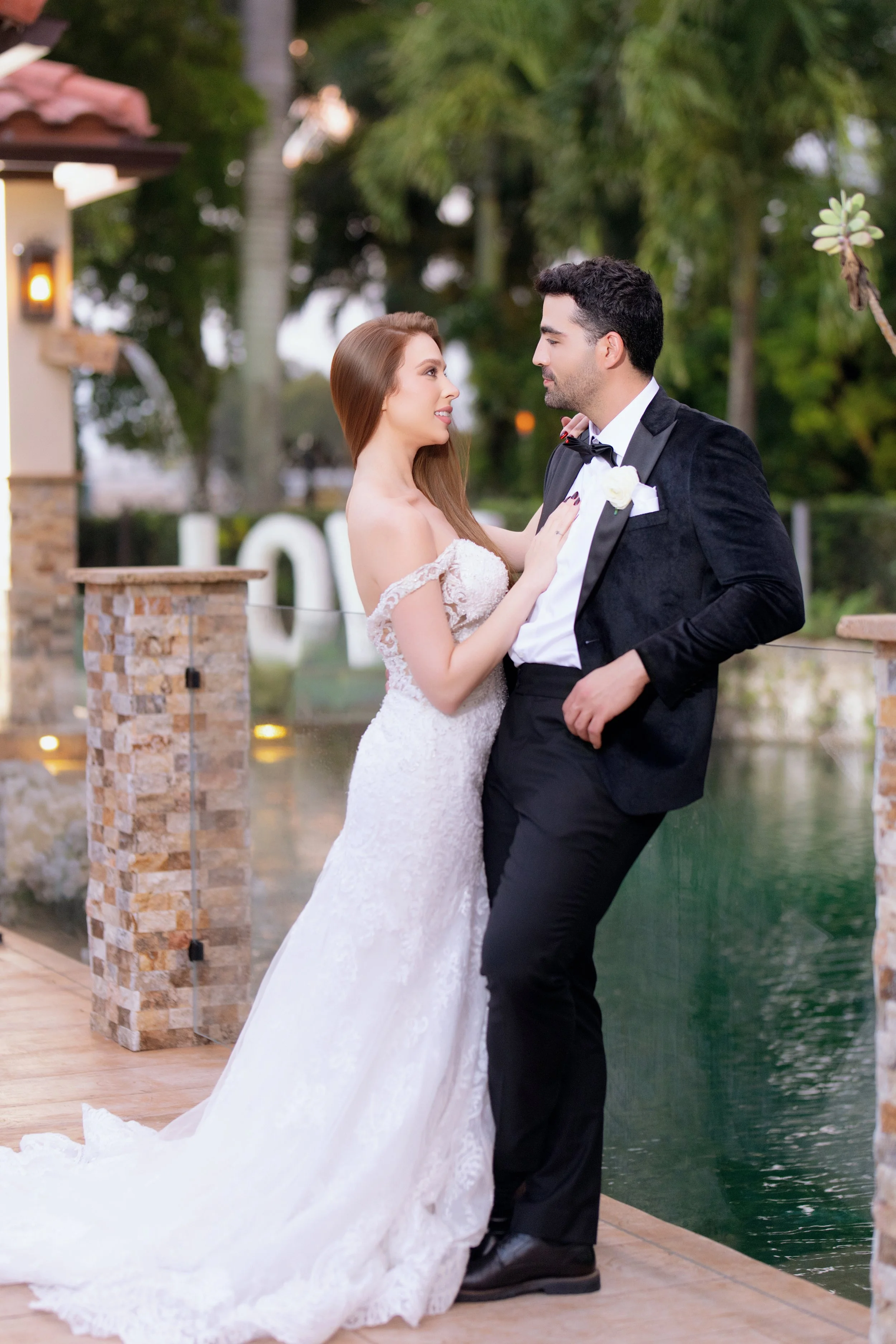 A bride and groom standing close together on a wooden dock by a water feature, gazing into each other's eyes, during their wedding celebration. The bride is in a white lace wedding gown with off-shoulder sleeves, and the groom is in a black tuxedo wi