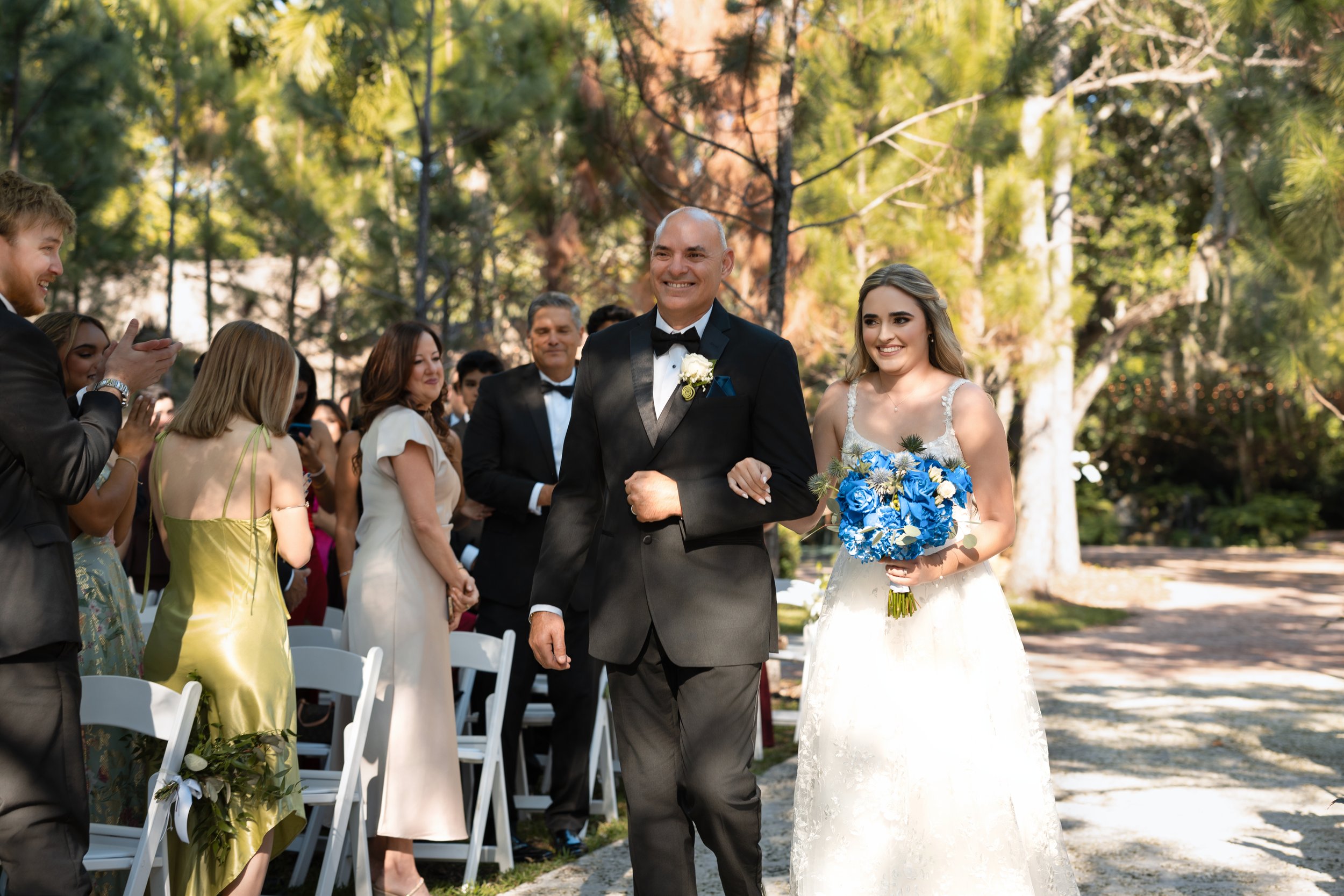 A bride and a man, likely her father, walking down an outdoor aisle at a wedding ceremony. The bride is holding a bouquet of blue flowers and smiling, while the man is dressed in a black tuxedo with a white boutonniere. Guests are standing and clappi