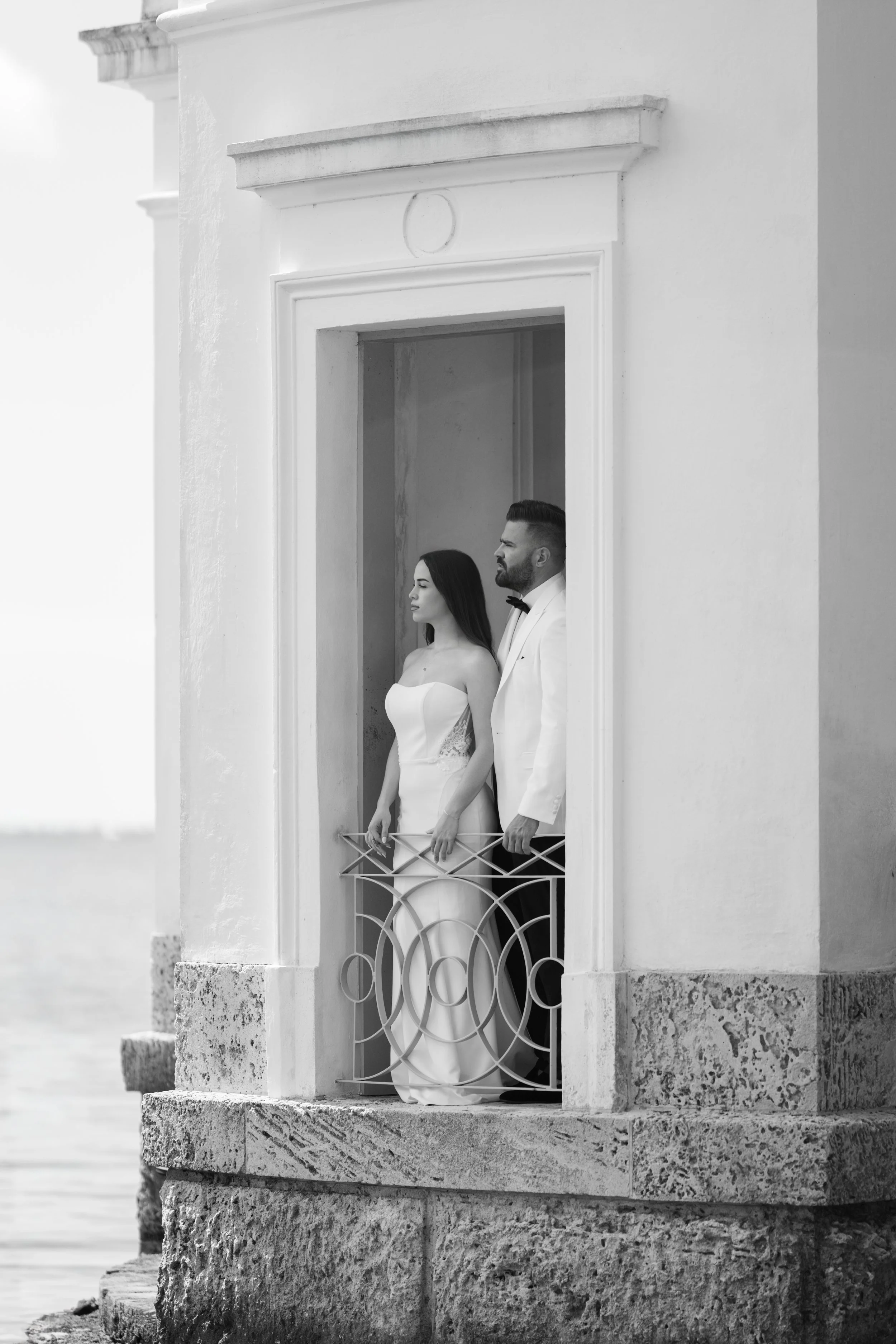 Black and white photo of a bride and groom standing on a small balcony outside a building, overlooking a body of water.