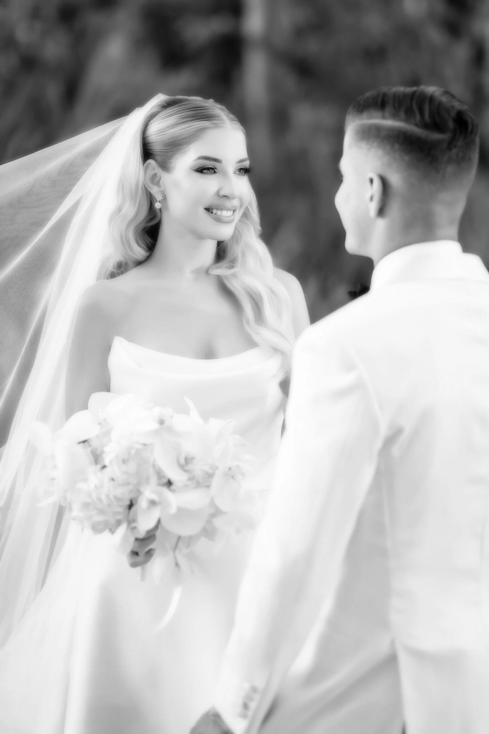 A black and white photograph of a bride and groom on their wedding day. The bride is smiling and holding a bouquet of flowers, wearing a strapless wedding dress and veil, with wavy hair and earrings. The groom is seen from the back, wearing a tuxedo,