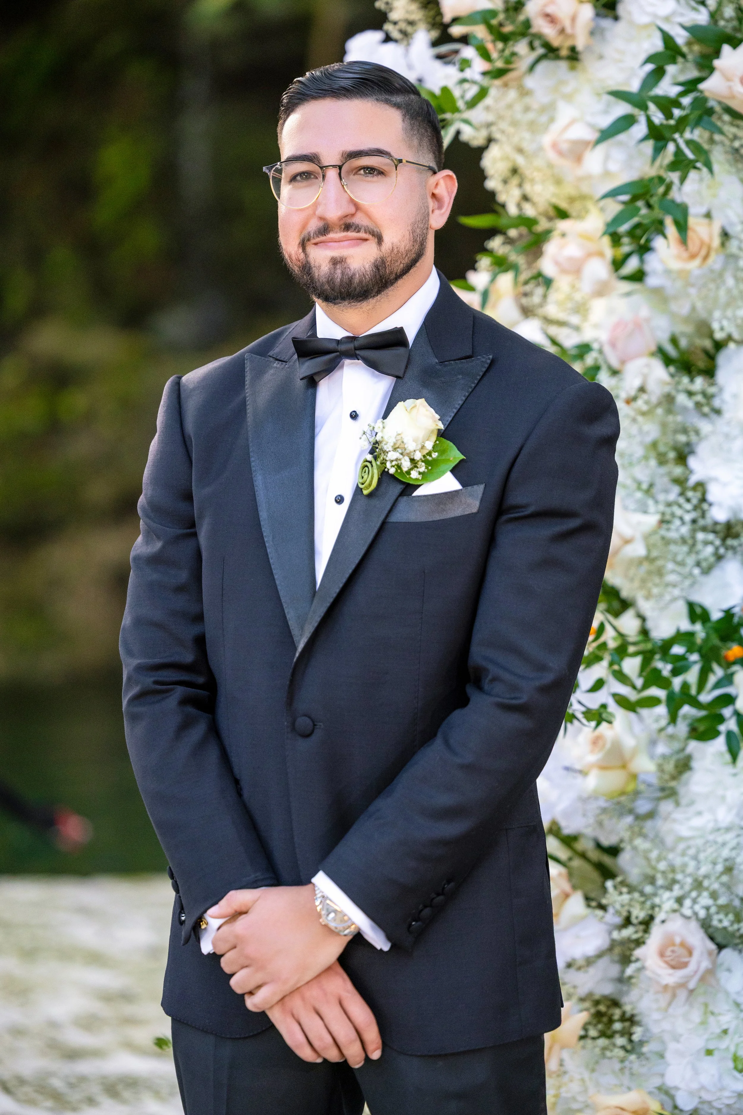 A groom in a black tuxedo with a black bow tie, glasses, and a boutonniere, standing in front of a floral backdrop at a wedding.