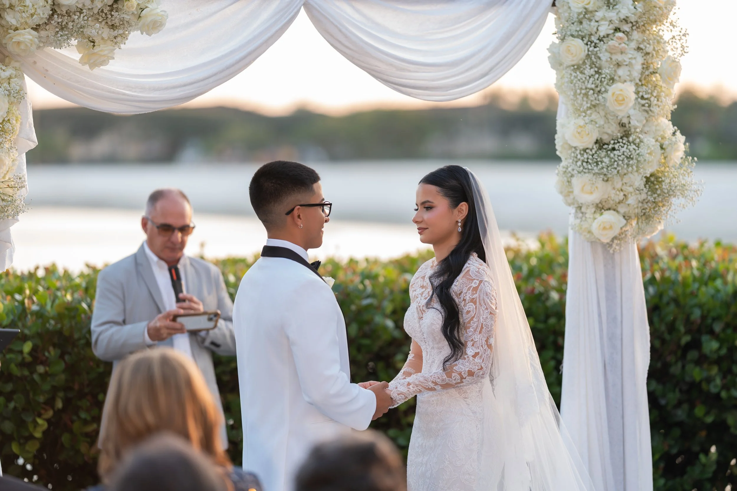 A couple getting married outdoors by a body of water, holding hands, with an officiant in the background, under a floral arch with white roses and drapery, during sunset.