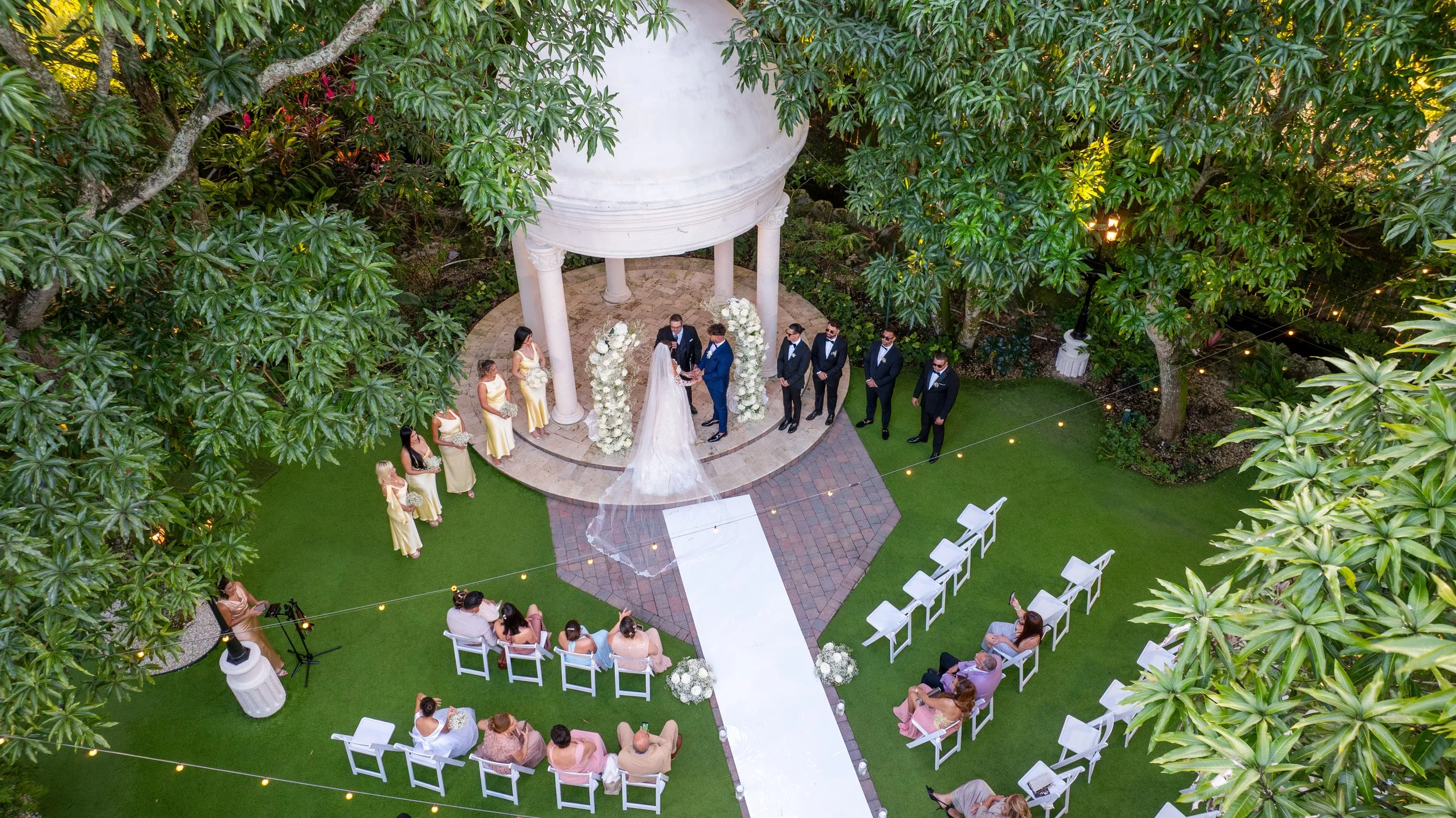 An outdoor wedding ceremony in a garden, with a bride and groom exchanging vows under a white gazebo surrounded by white floral arrangements. Guests seated on white chairs watch the ceremony, and bridesmaids and groomsmen stand on either side.