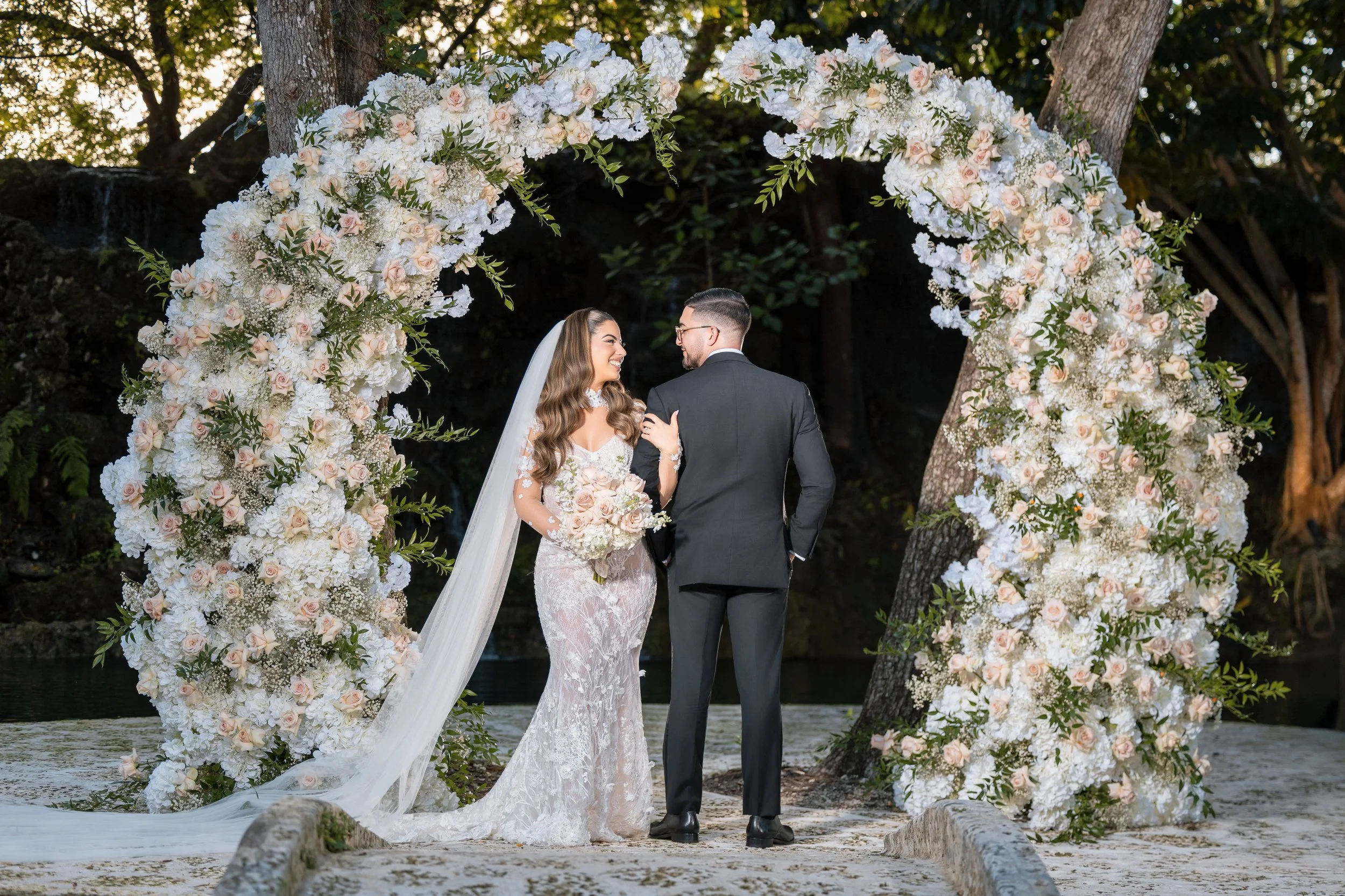 Bride and groom standing under a floral arch during their wedding ceremony on a sandy beach with trees in the background.