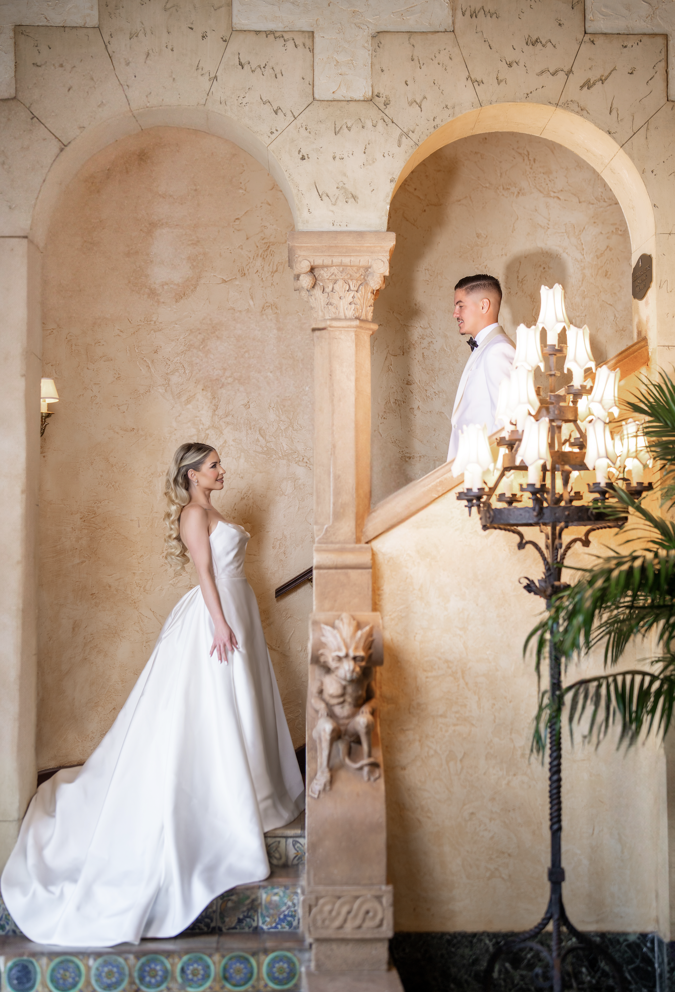 A bride in a white wedding gown standing on stairs looking at a groom in a white tuxedo on a staircase in an elegant, warm-toned interior with archways and ornate decor.