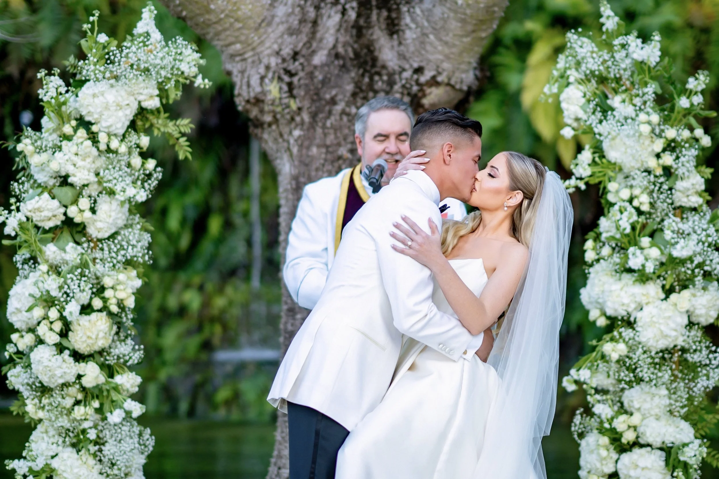 A couple in wedding attire sharing a kiss at their outdoor wedding ceremony, with an officiant standing behind them and floral arrangements on either side.