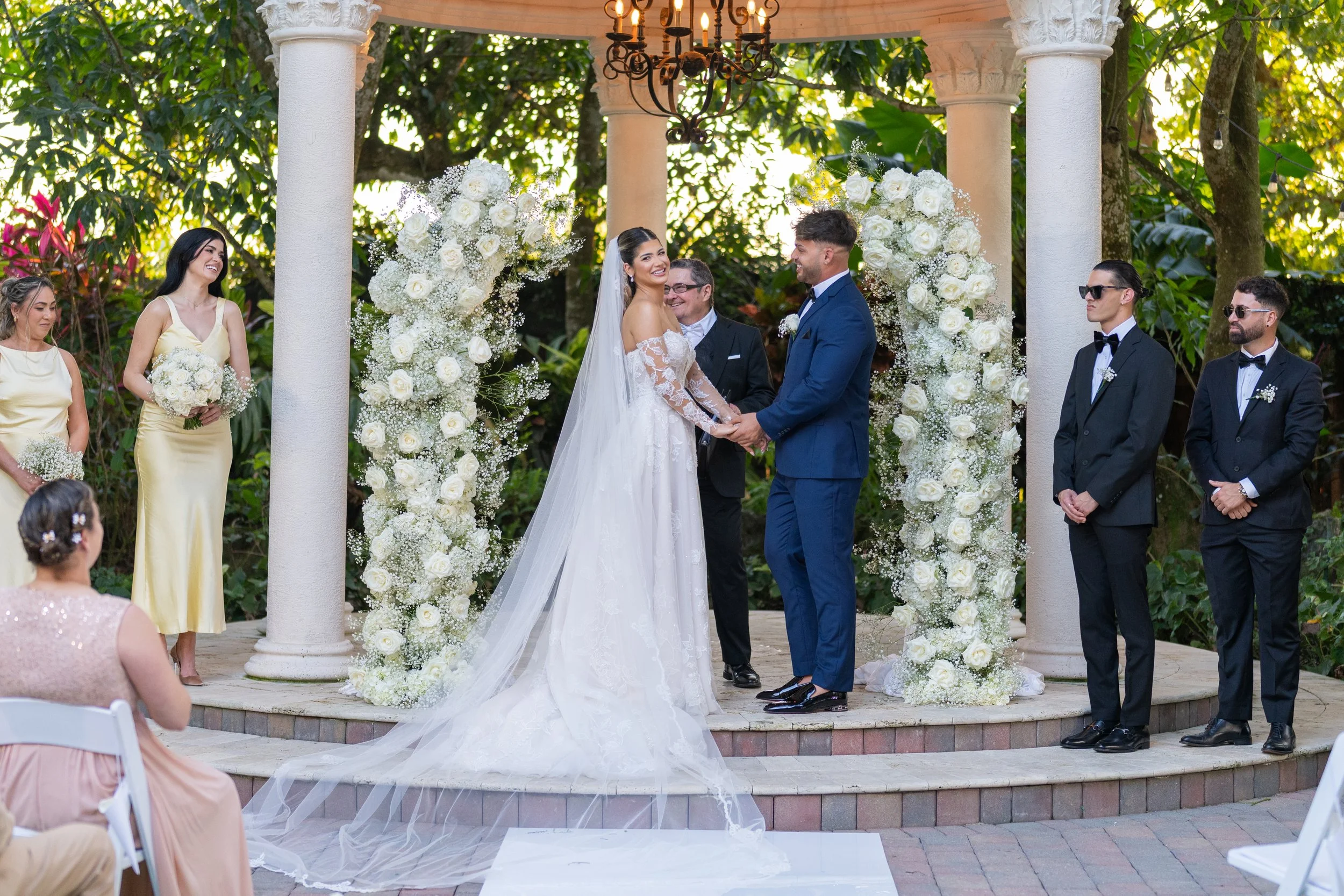 A wedding ceremony with a bride and groom holding hands in front of an outdoor floral arch, surrounded by bridesmaids and groomsmen, during daytime.