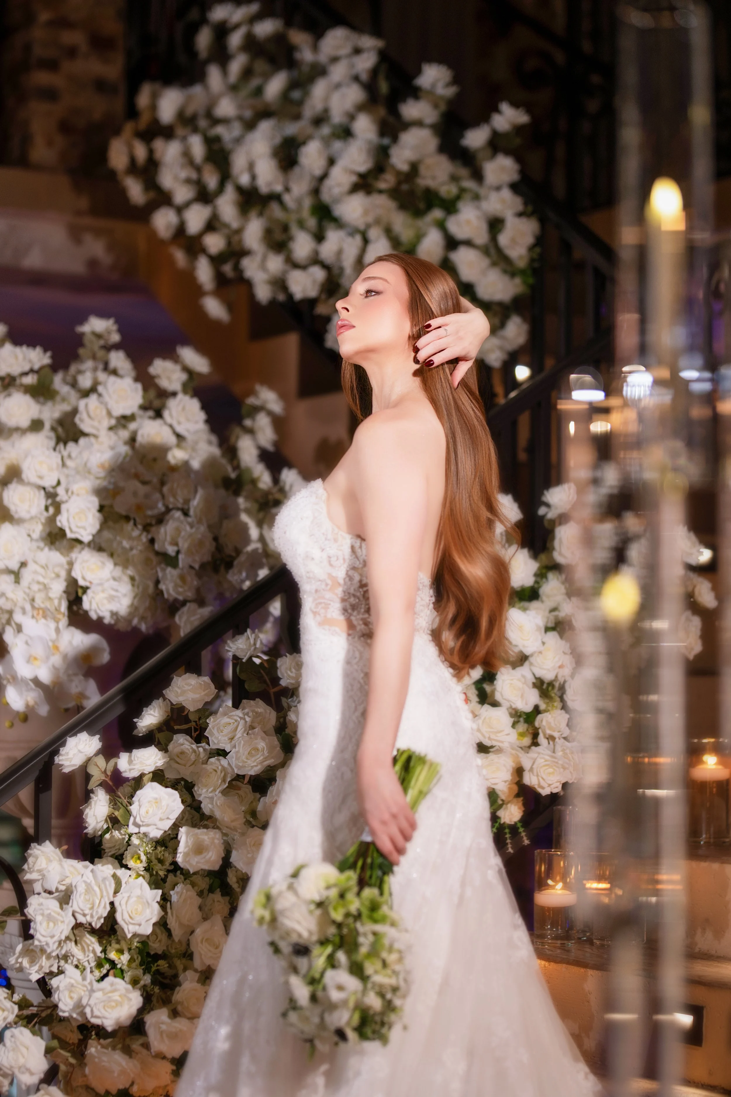 A bride in a white wedding dress holding a bouquet of white roses, standing near a staircase decorated with white roses.