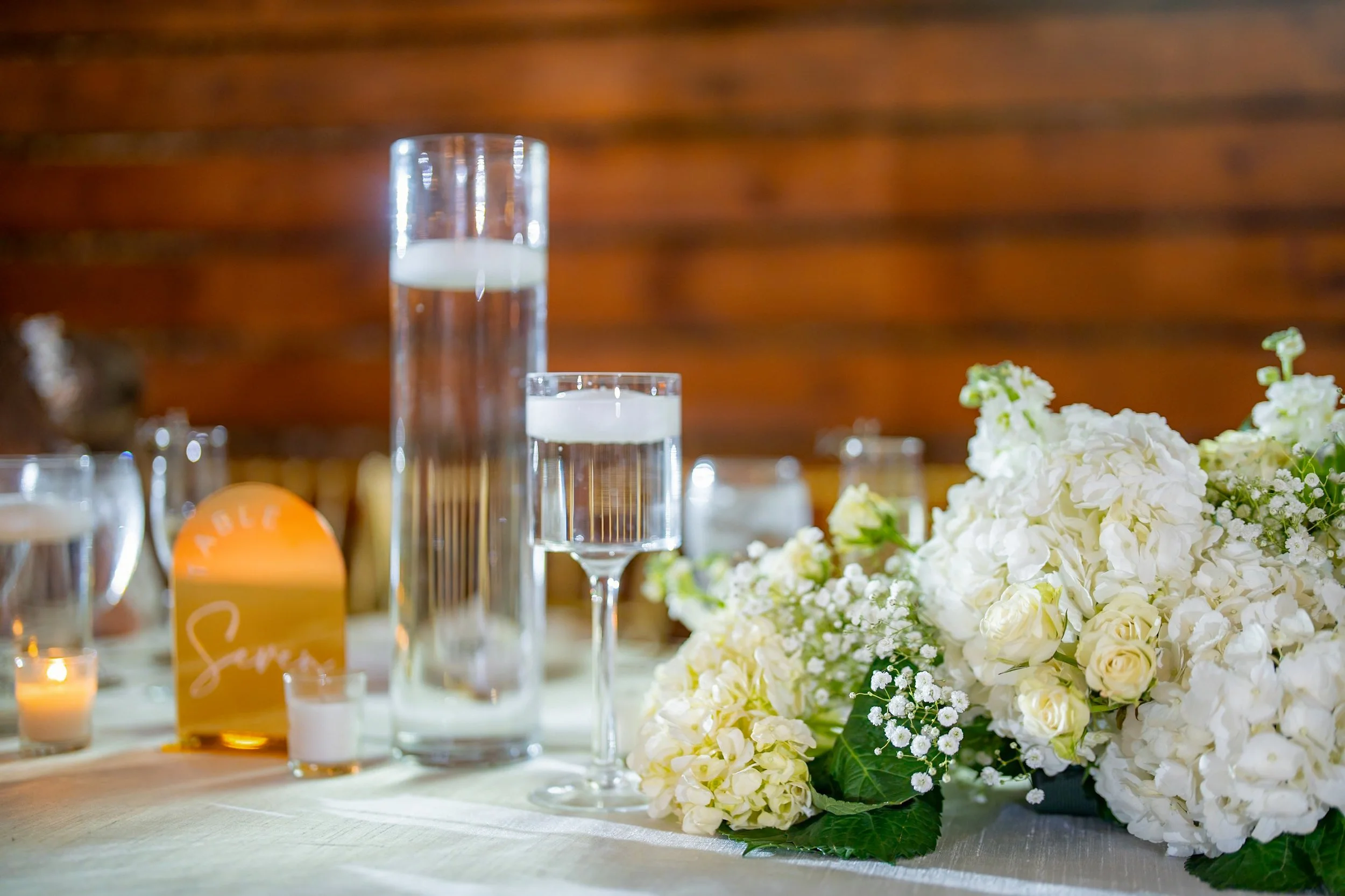 A wedding reception table with floral arrangements, glassware, and candles, with a wooden wall in the background.