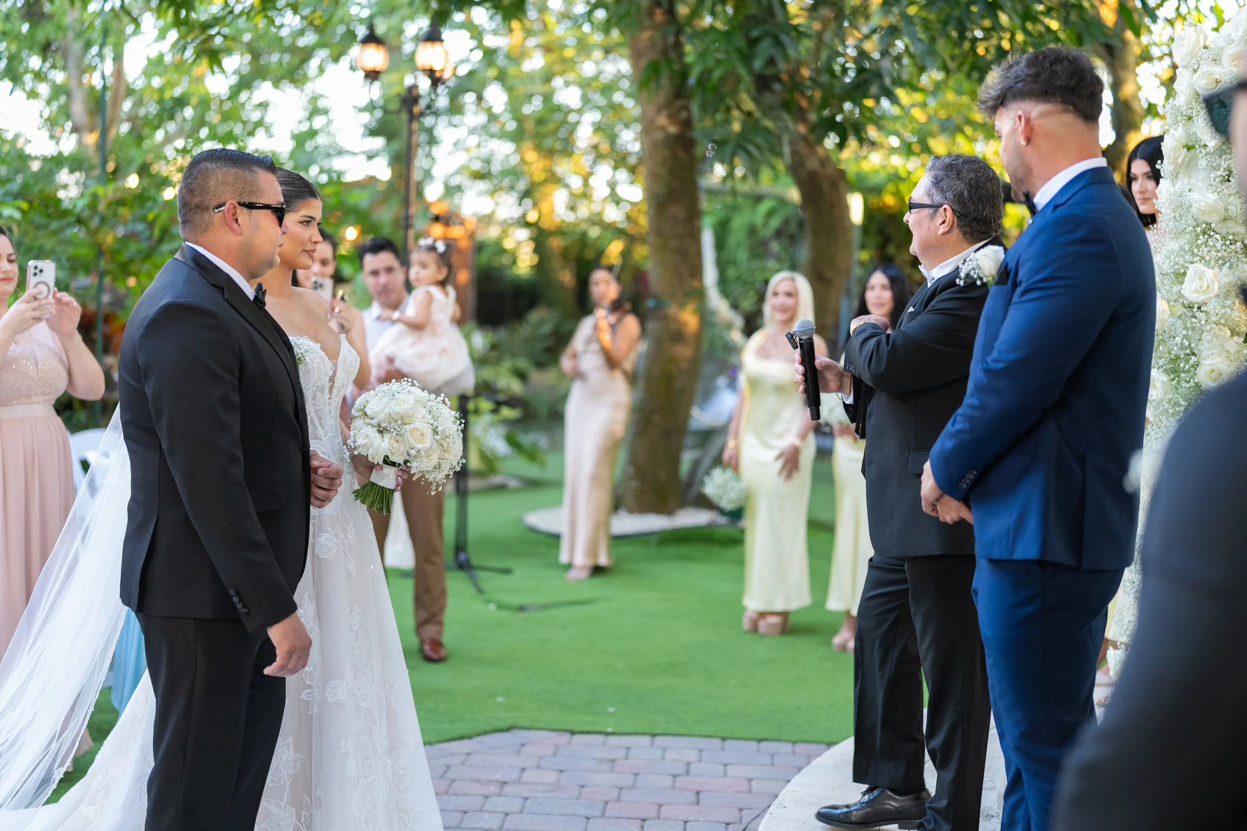 Wedding ceremony outdoors with bride and groom facing officiant, surrounded by guests and greenery.