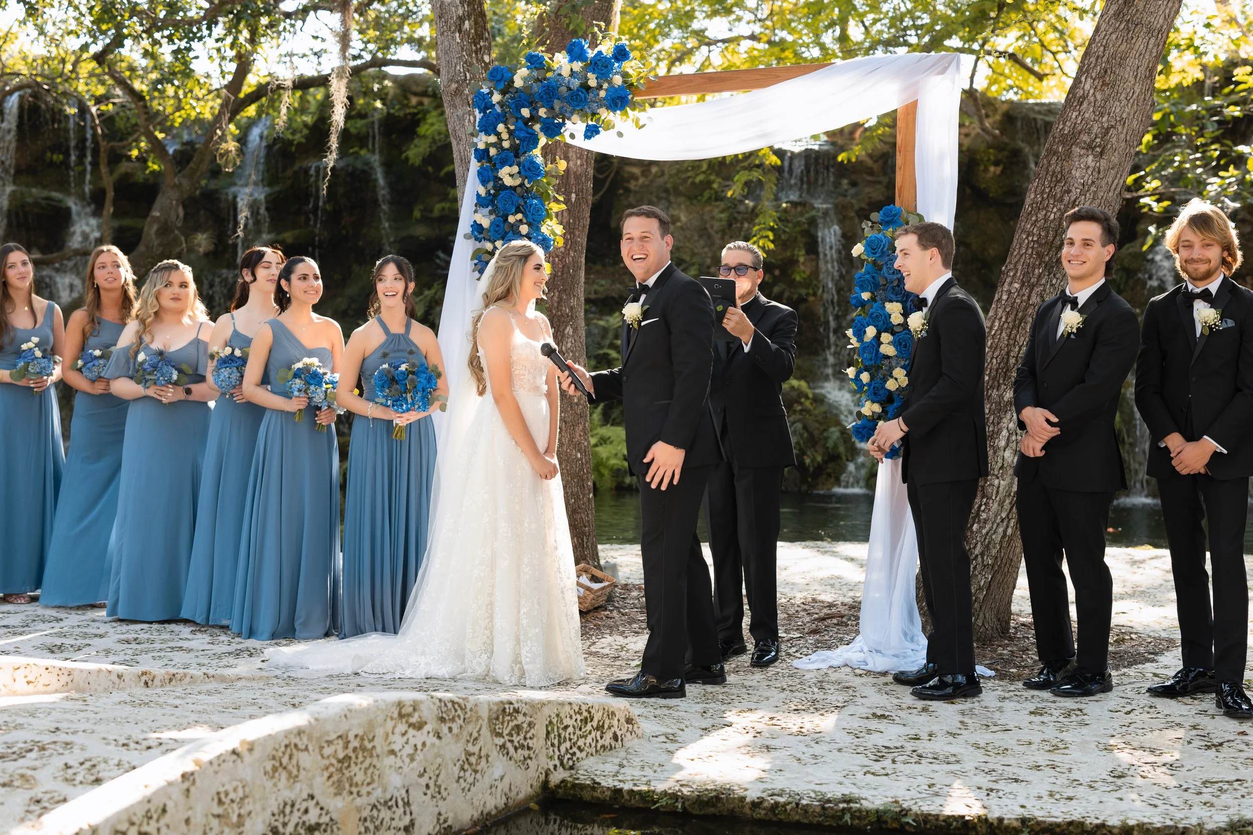 A wedding ceremony taking place outdoors near a waterfall, with the bride and groom exchanging vows, surrounded by bridesmaids in blue dresses and groomsmen in black tuxedos.