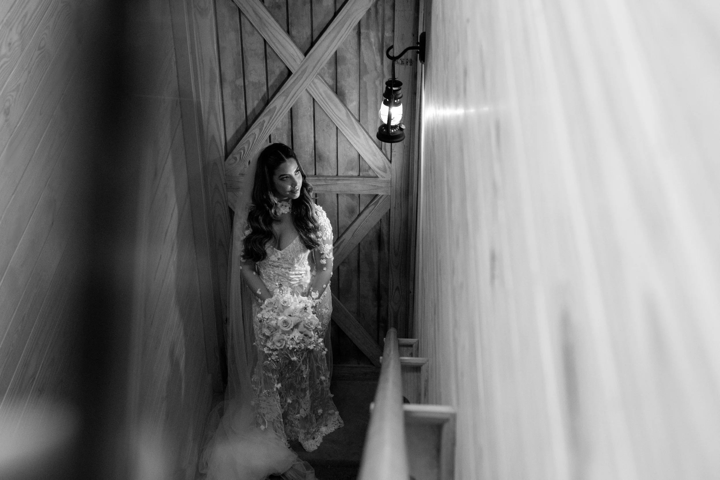A woman in a wedding dress holding a bouquet of roses, standing in a narrow wooden staircase enclosed by wooden walls, illuminated by a hanging lantern.