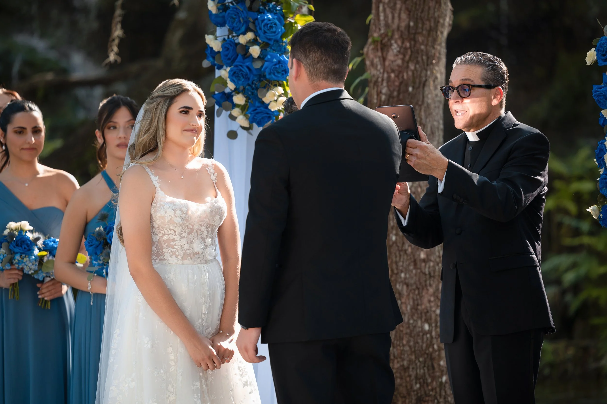 A couple gets married outdoors with a priest officiating, surrounded by bridesmaids in blue dresses holding bouquets and adorned with blue and white floral arrangements, during a daytime wedding ceremony.