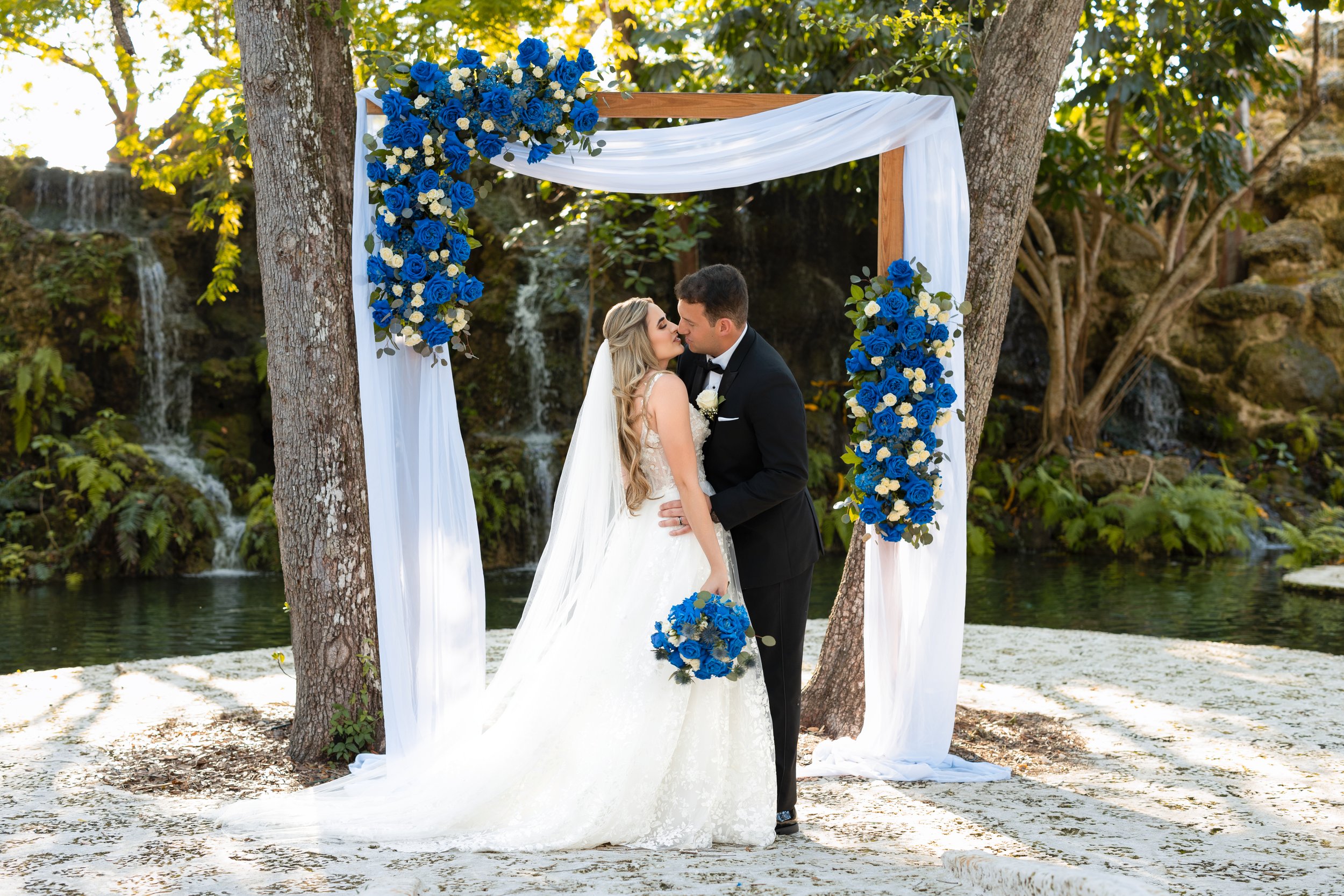 A bride and groom share a kiss under a white-and-blue floral wedding arch outdoors near water and trees.