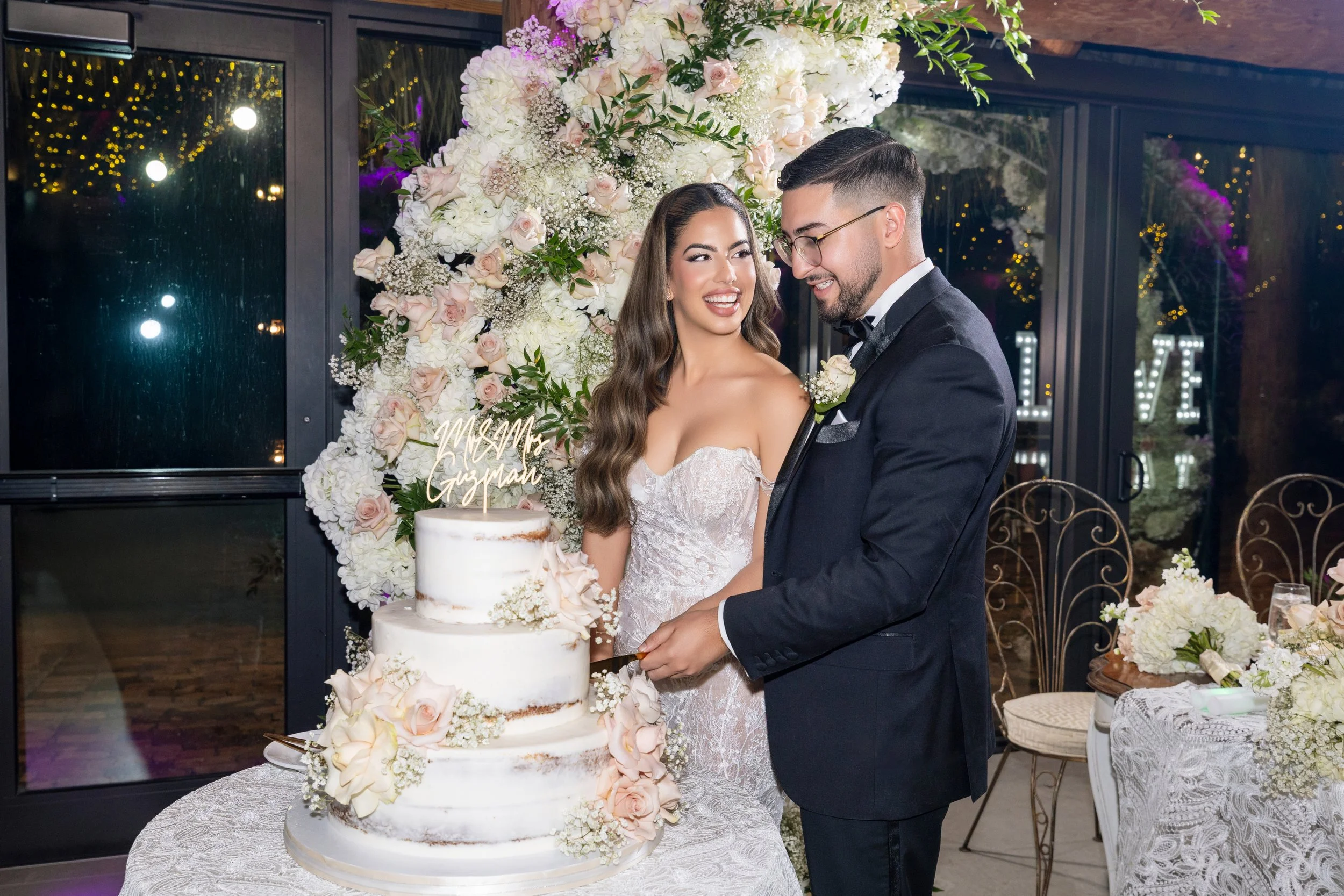 A newlywed couple, a woman in a white wedding dress and a man in a black tuxedo, sharing a moment by their wedding cake decorated with flowers, inside a wedding reception venue with floral arrangements and lights in the background.