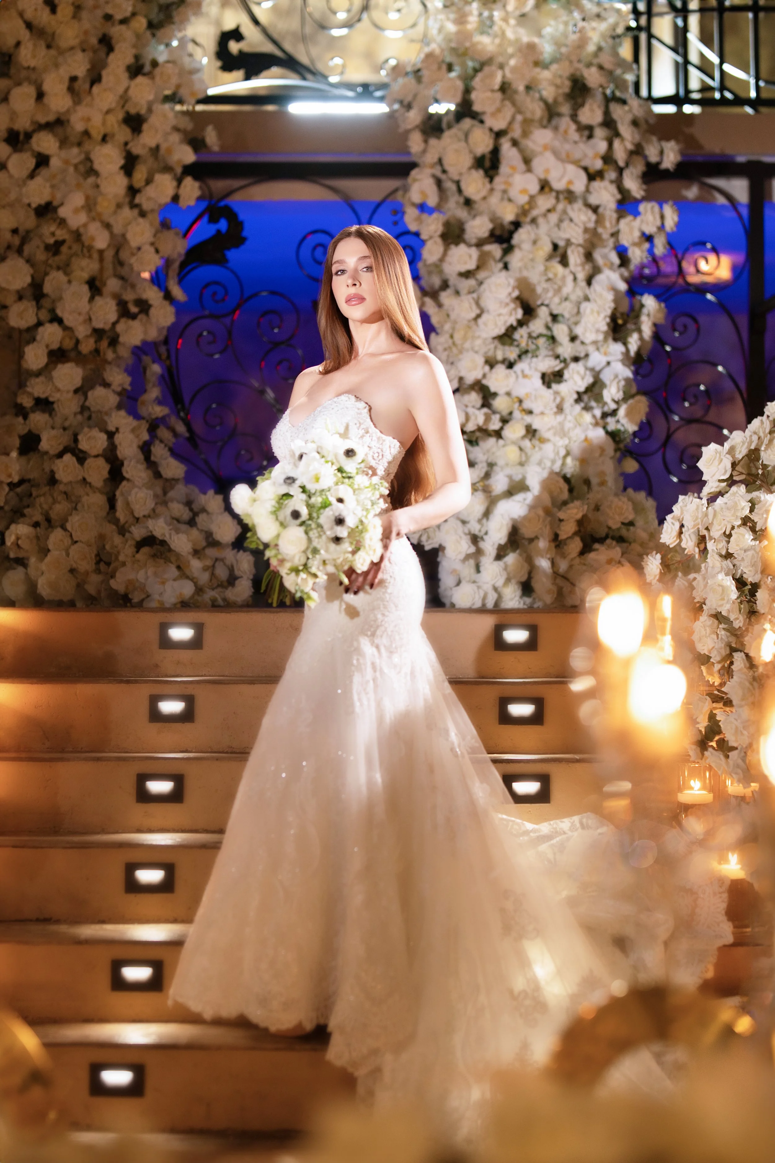 A bride standing on a staircase holding a bouquet of white flowers, surrounded by a floral arch of white roses and candles, in a decorated wedding setting with purple and blue lighting.