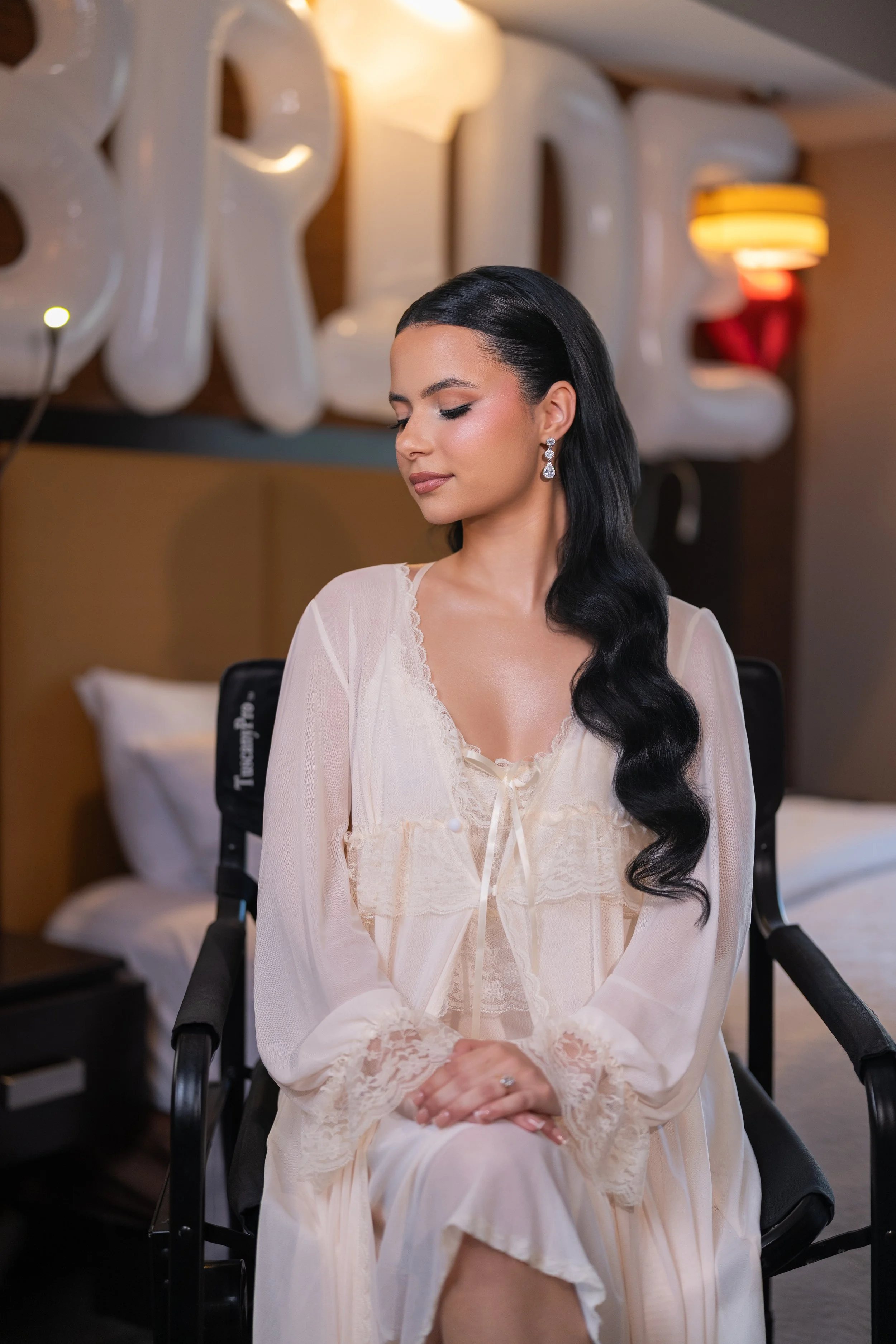 A woman with long black wavy hair, dressed in a cream-colored lace nightgown, sits silently with her eyes closed in a hotel room. Behind her, large white balloon letters spell out "PRIDE" and a lamp emits a warm glow.