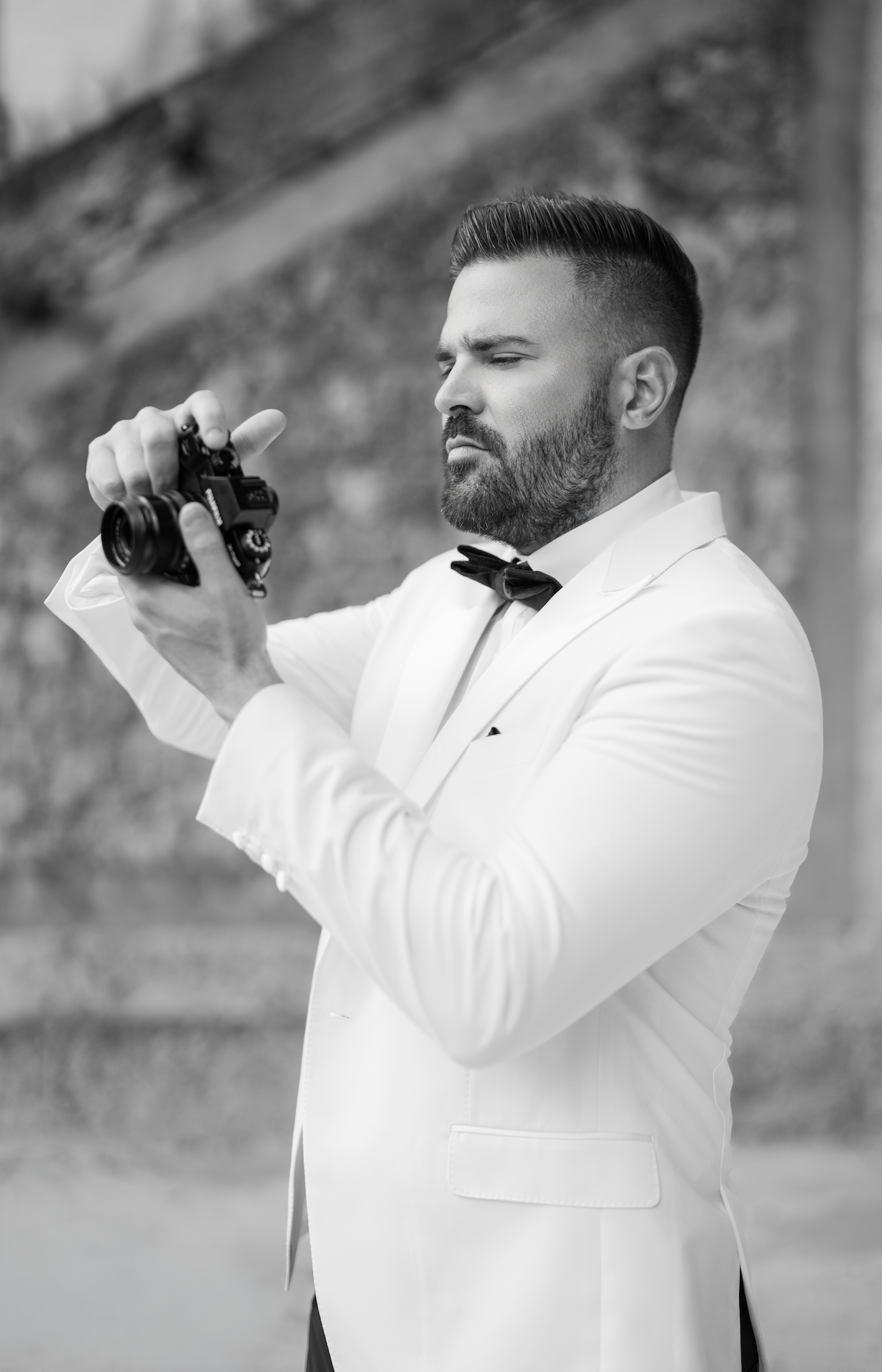 A man dressed in a white tuxedo with a black bow tie is holding a camera and looking at it thoughtfully. The background is blurred, showing an outdoor setting.