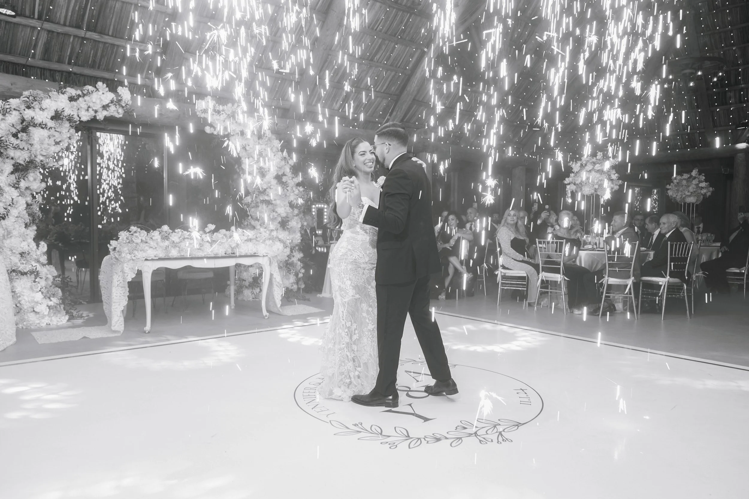 A bride and groom dancing on a circular dance floor during their wedding reception, surrounded by sparklers and seated guests in a decorated venue.
