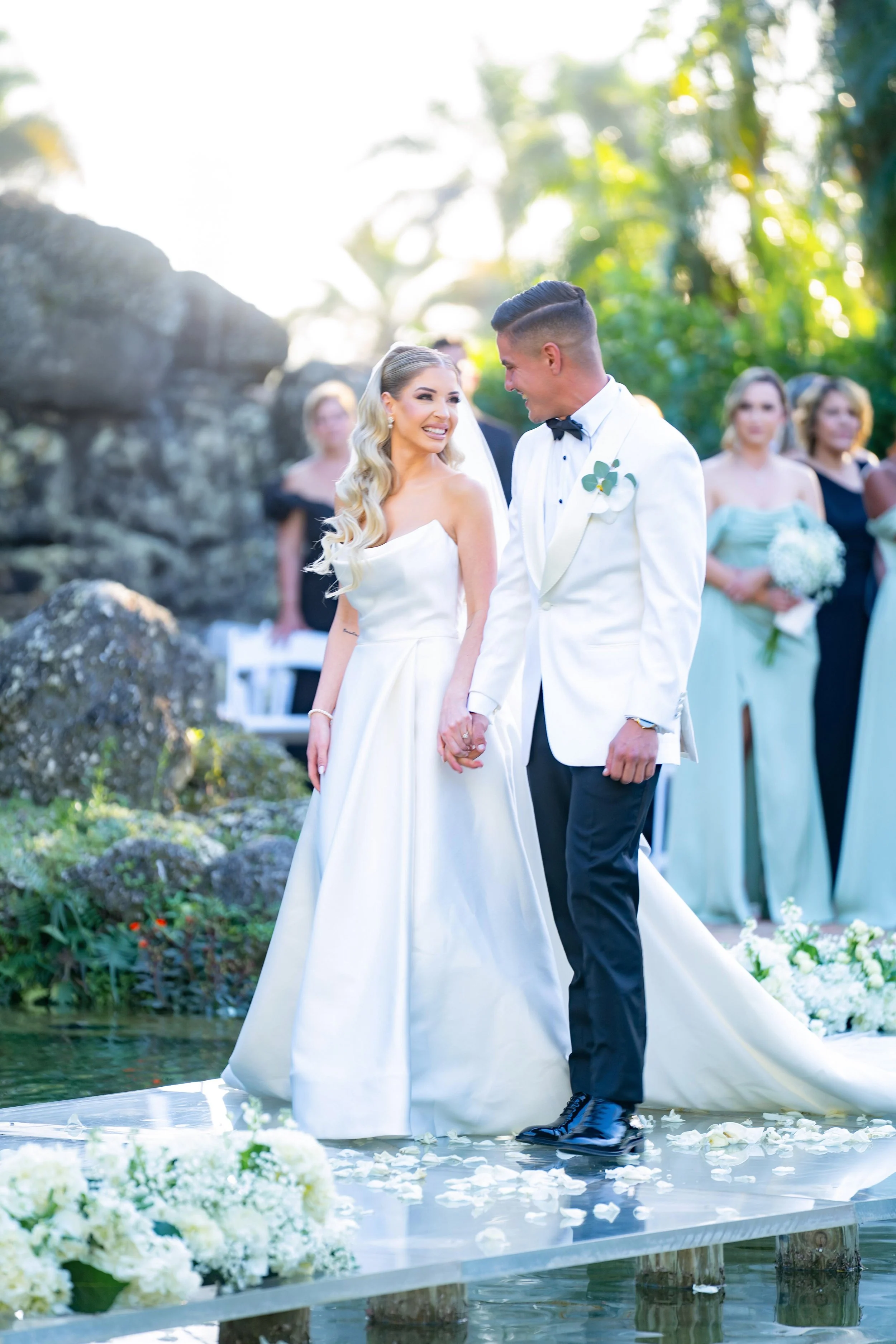 A bride and groom holding hands during an outdoor wedding ceremony on a platform over water. The bride wears a white wedding gown with a sweetheart neckline, and the groom wears a white tuxedo jacket with black pants and a bowtie. Bridesmaids and gue