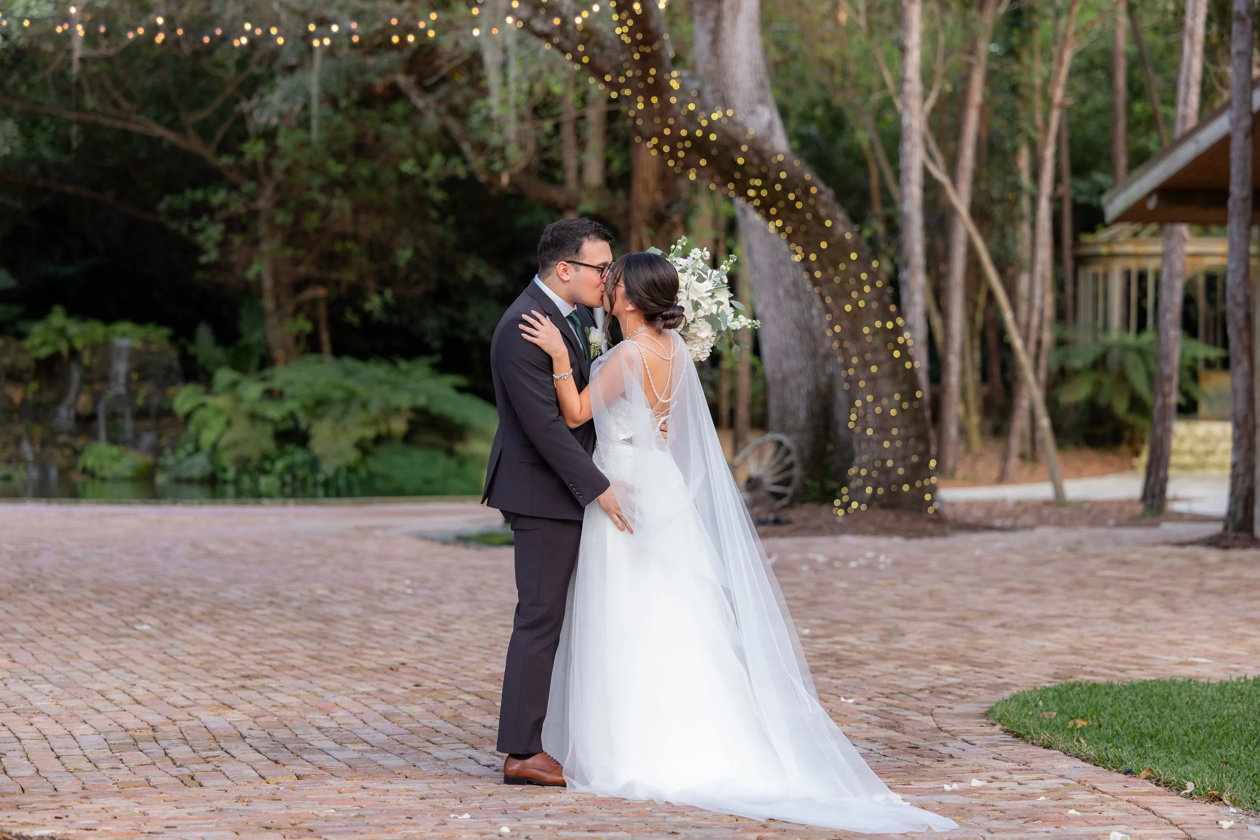 A bride and groom sharing a kiss on their wedding day outdoors, with trees and string lights in the background.