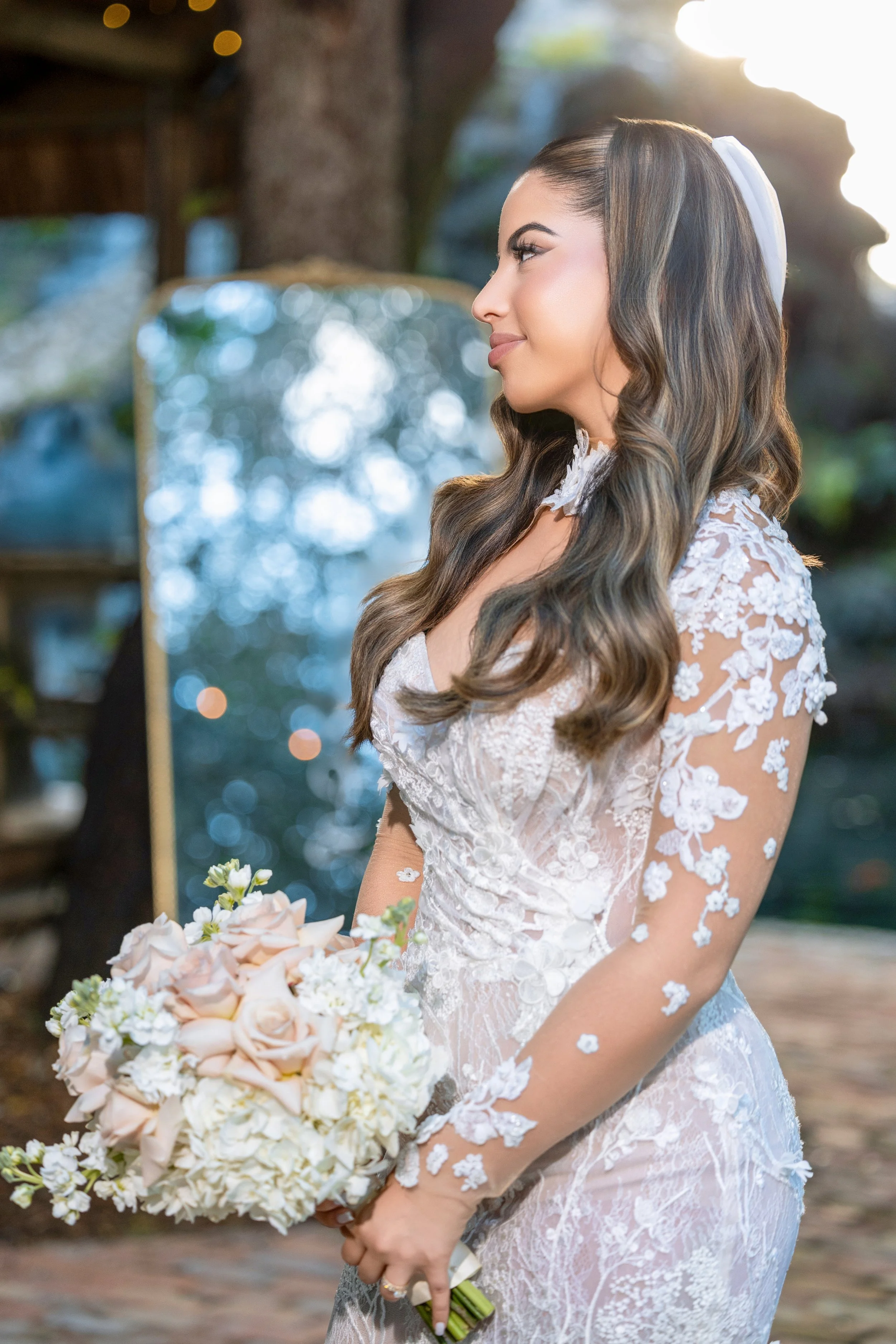 A bride with long, wavy brown hair holding a bouquet of pink and white roses and hydrangeas, wearing a lace wedding dress with floral appliqués and a white ribbon in her hair, standing outdoors near a tree with soft sunlight in the background.