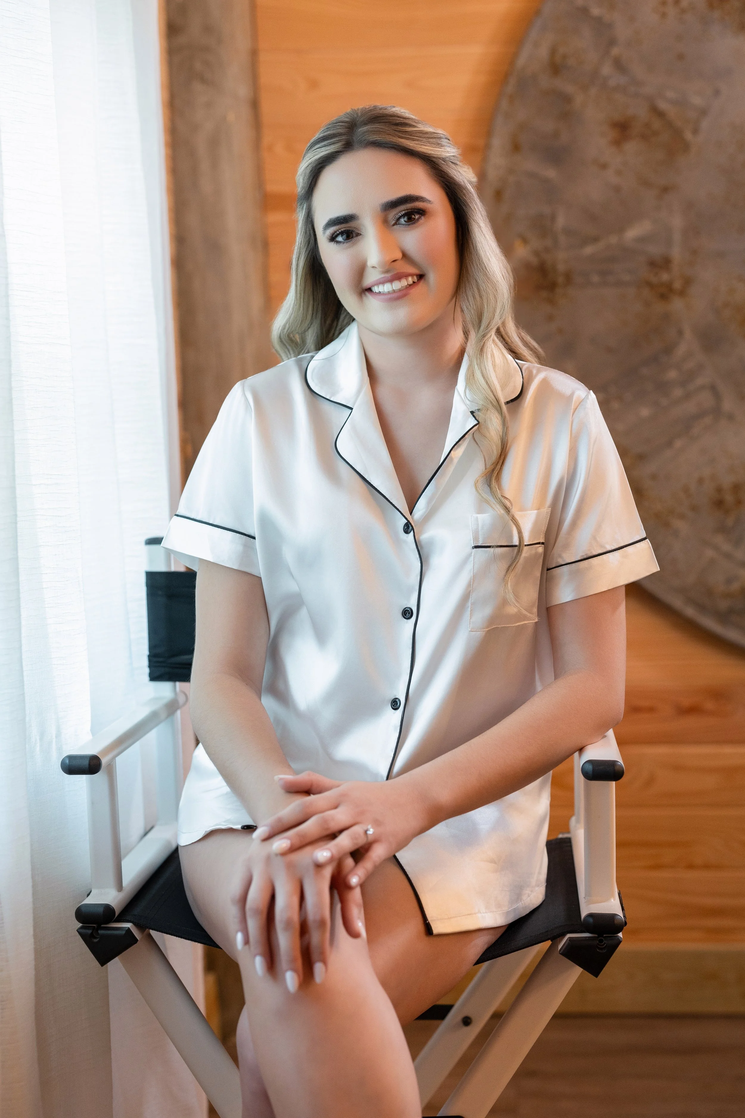 A young woman with blonde hair and fair skin sitting on a black folding chair indoors, wearing a cream-colored silk pajama top with black piping.