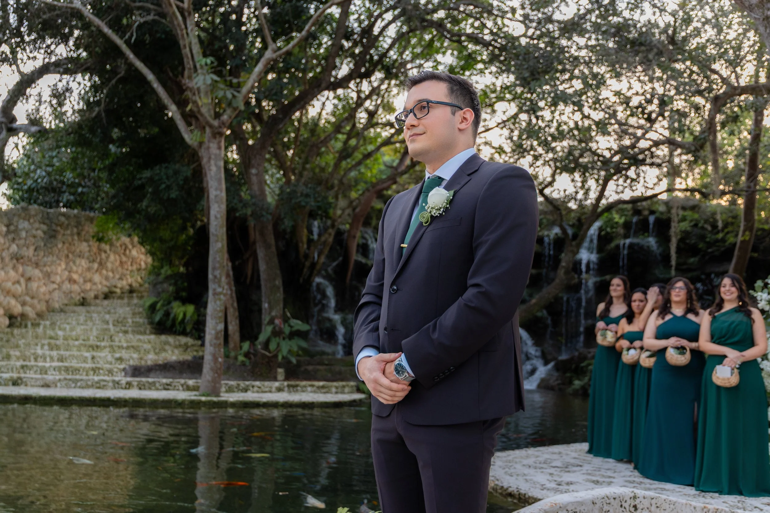 A groom in a black suit and glasses stands outdoors near a pond, with a waterfall and trees in the background, during a wedding ceremony. Bridesmaids in green dresses stand in the background holding baskets.