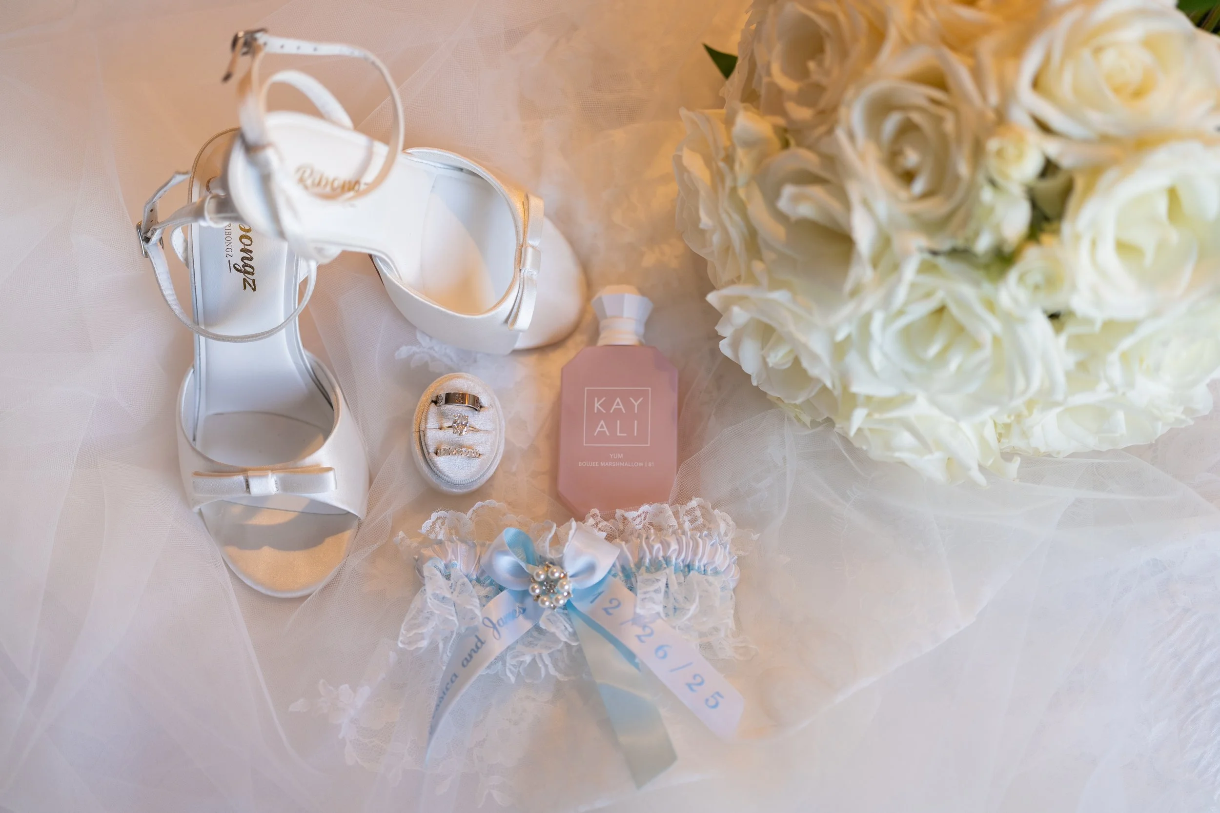 Wedding shoes, a ring box with rings, a bottle of perfume, a garter, and a bouquet of white roses arranged on a white surface.