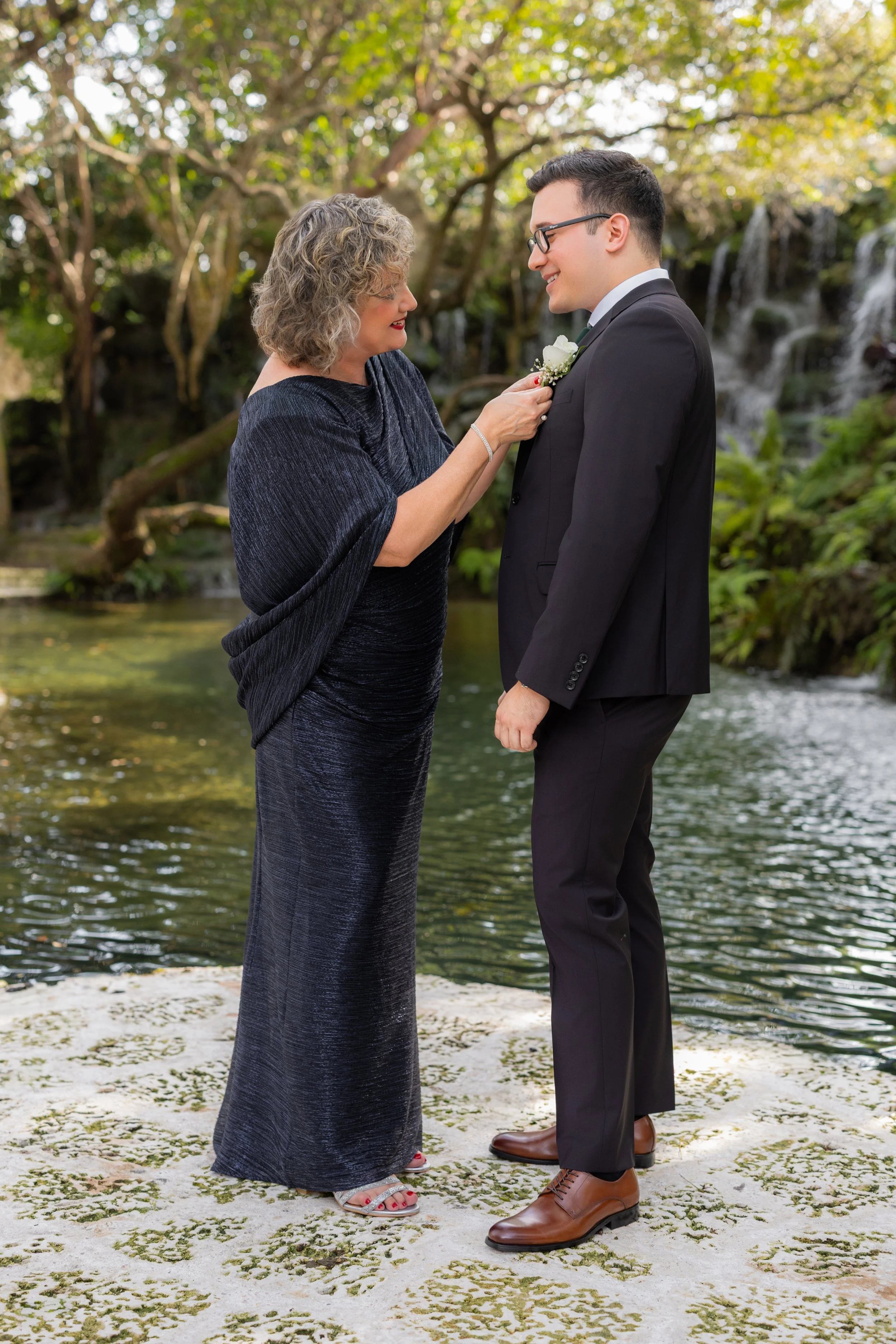 A woman in a dark blue dress is pinning a white boutonniere on a man in a black suit, who is smiling. They are standing outdoors by a waterfall and surrounded by lush trees.