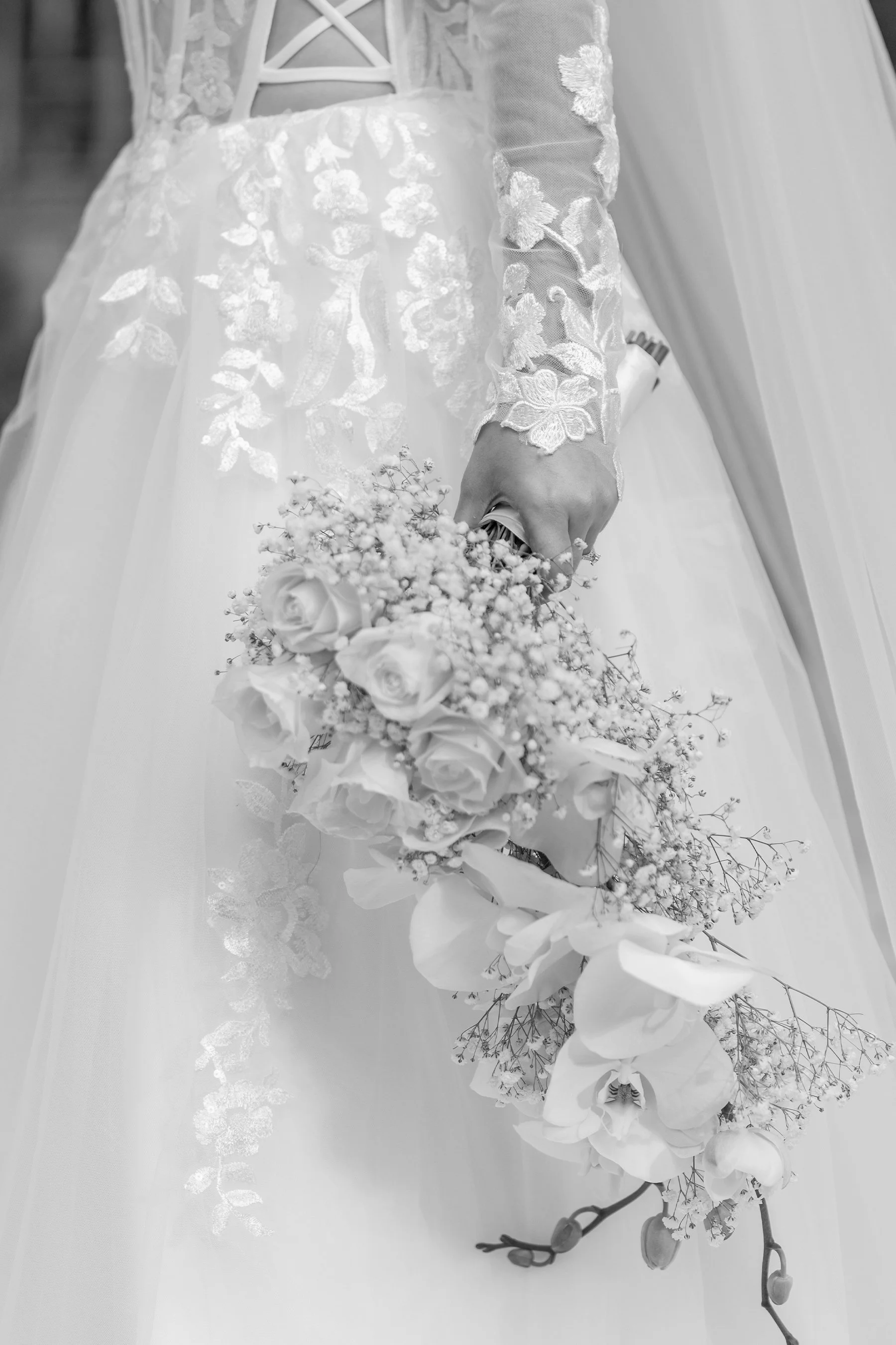 Close-up of a bride holding a bouquet of roses, baby's breath, and orchids, wearing a wedding dress with lace sleeves and floral embroidery.