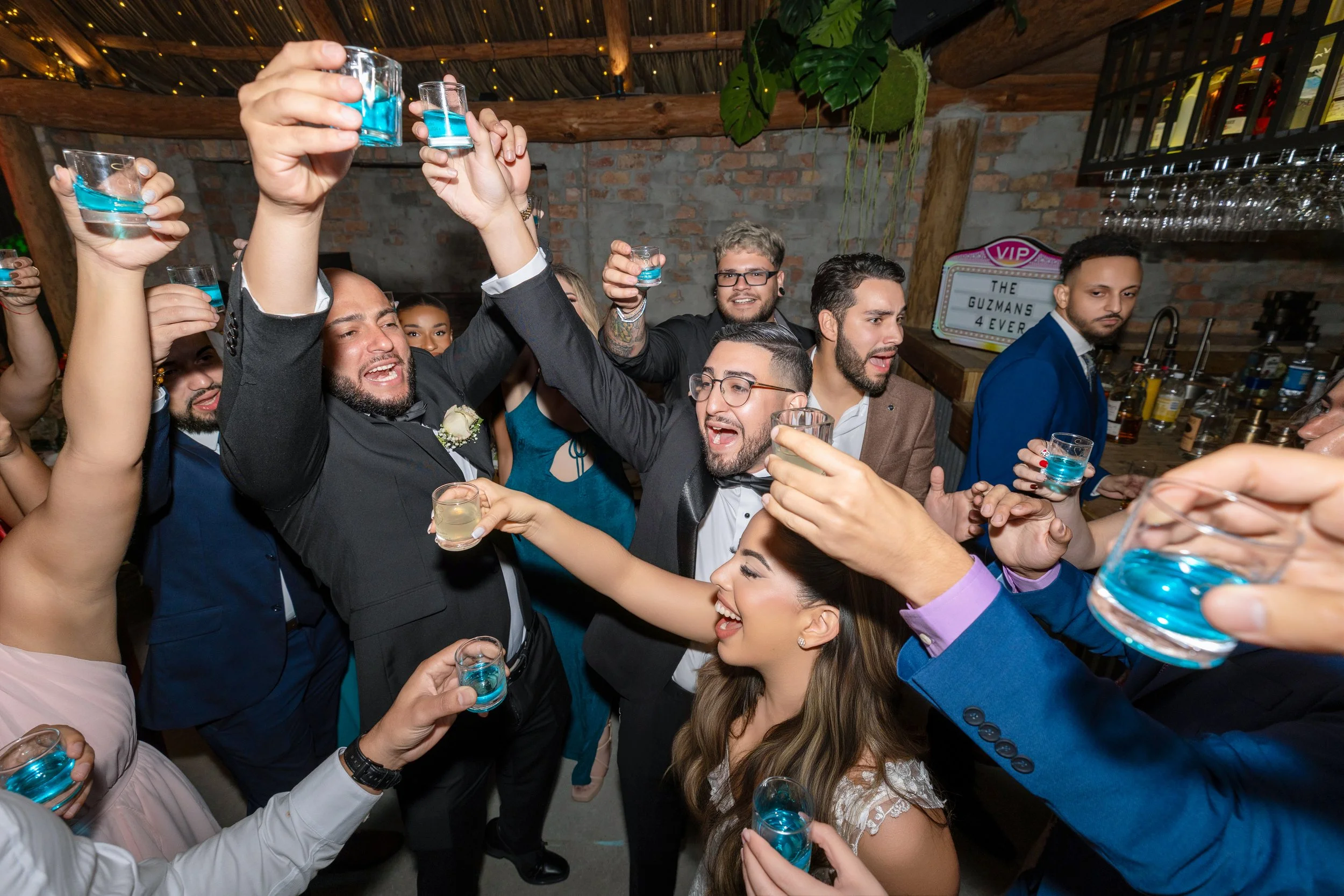 Group of people at a celebration party raising glasses with blue drinks, smiling and enjoying the moment. Indoor setting with brick wall, greenery, and a bar in the background.