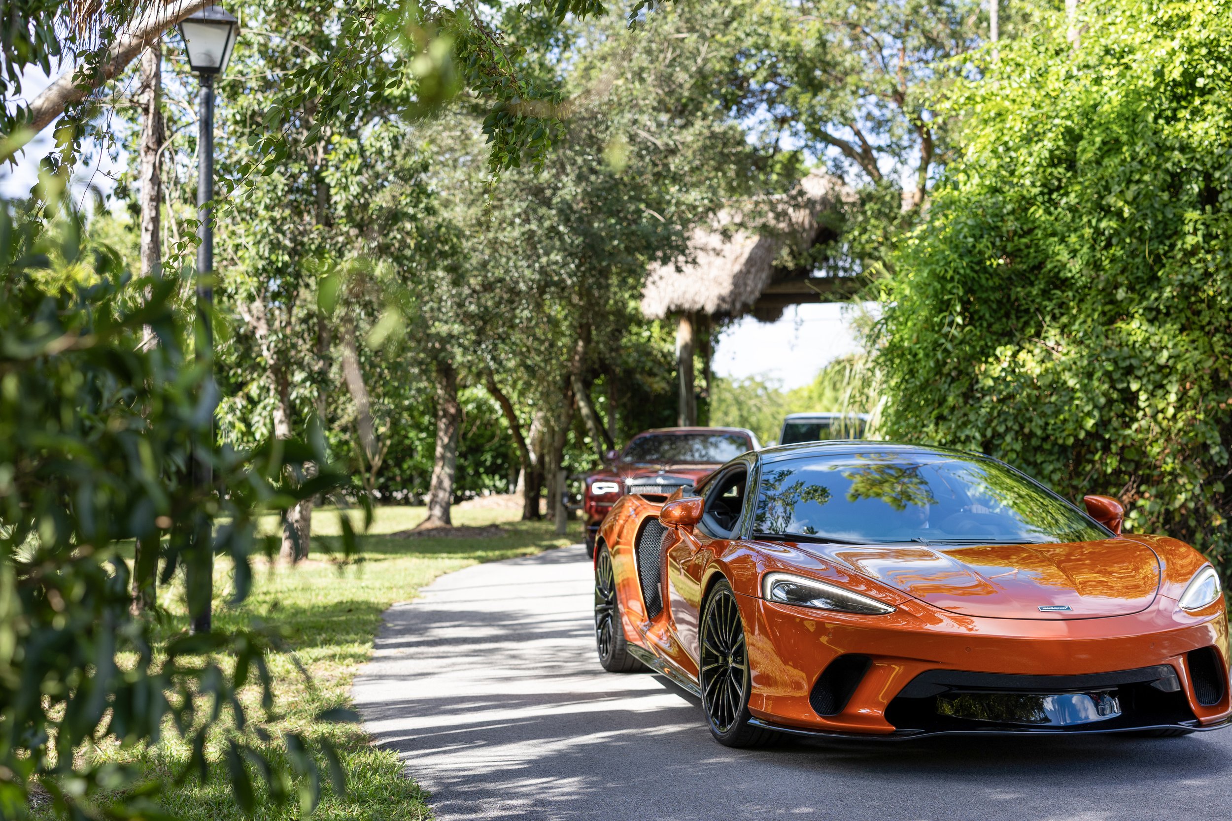 Orange sports car parked on a driveway surrounded by trees and greenery, with a thatched-roof structure and two other vehicles in the background.
