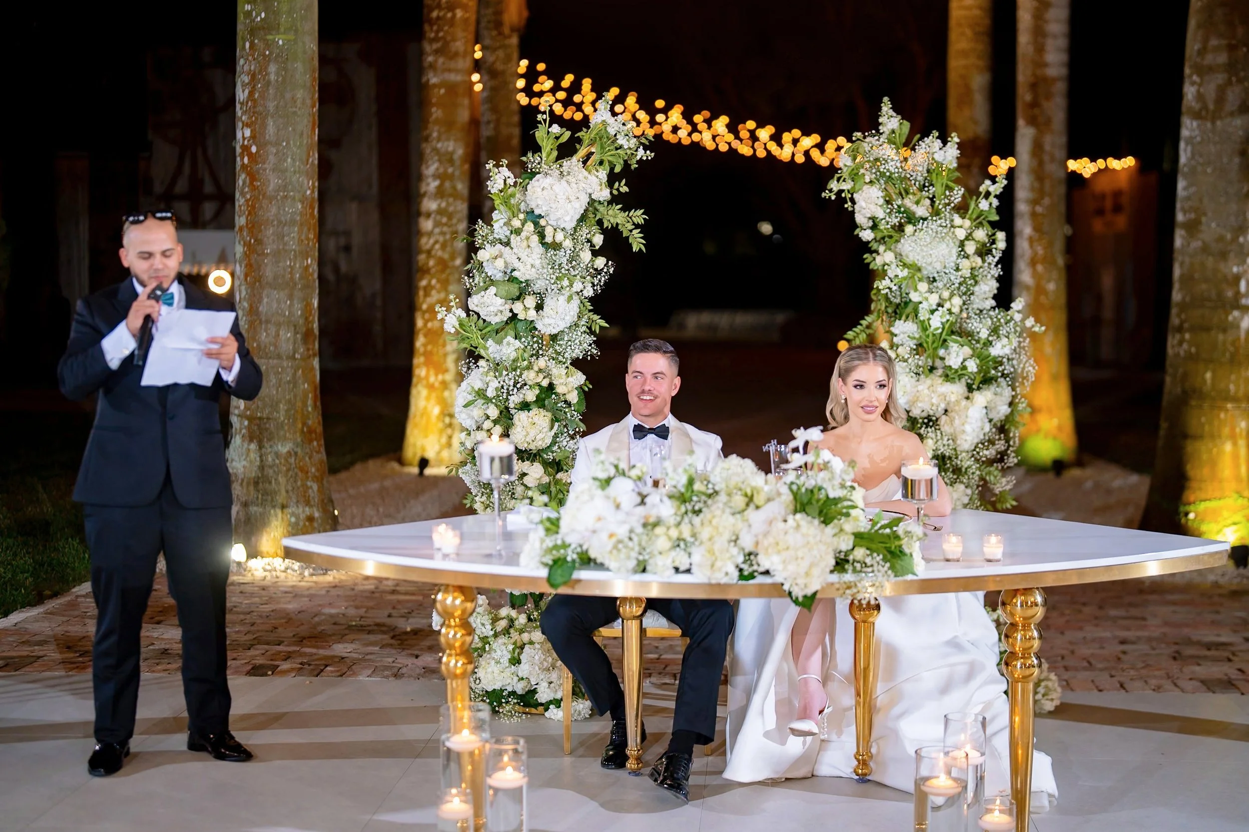 A wedding reception with a bride and groom sitting at a decorated table with white flowers and candles, outdoors at night, with a person giving a speech.