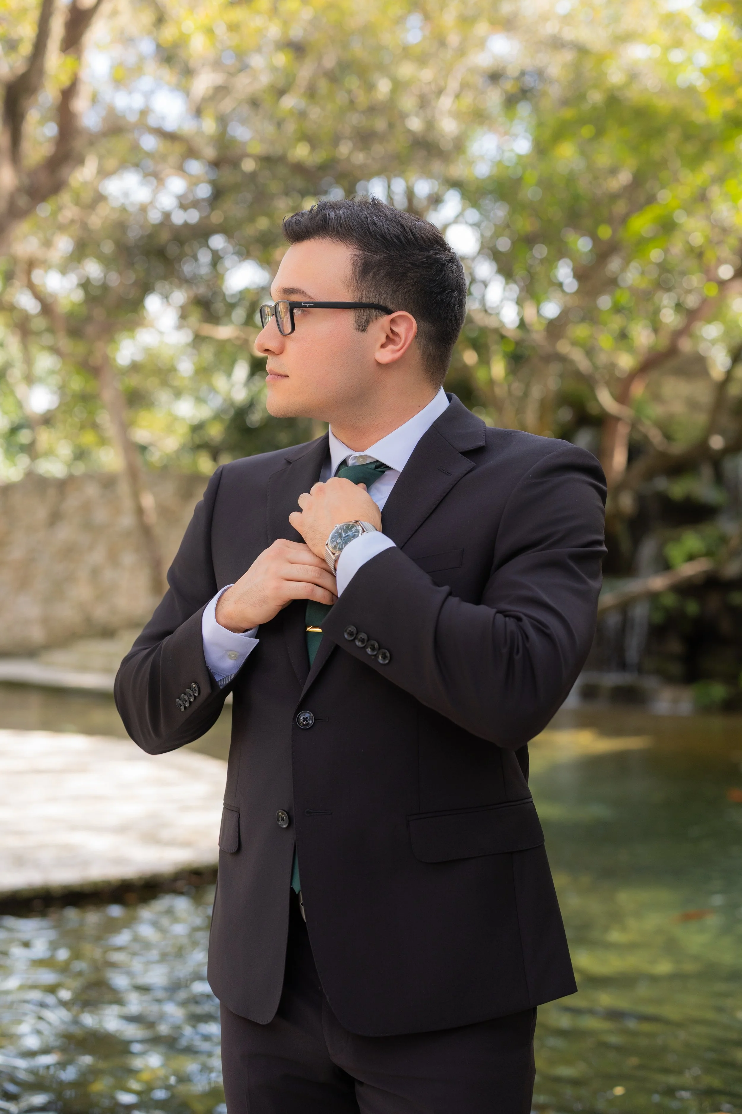 A man in a black suit, white shirt, and tie adjusting his cufflink outdoors near a water feature with trees in the background.