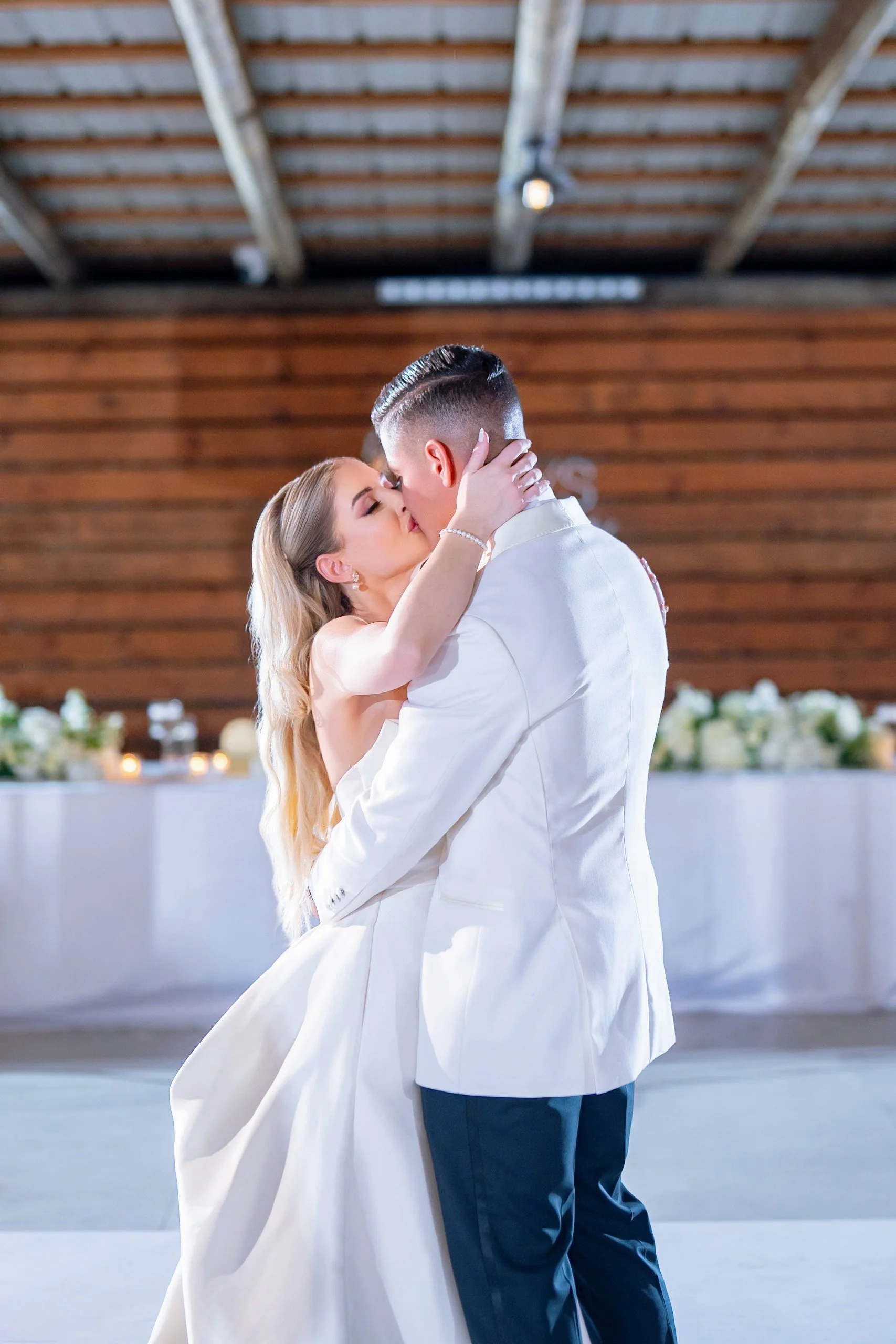 A bride and groom are dancing and kissing at their wedding reception, with the bride in a white gown and the groom in a white suit jacket.