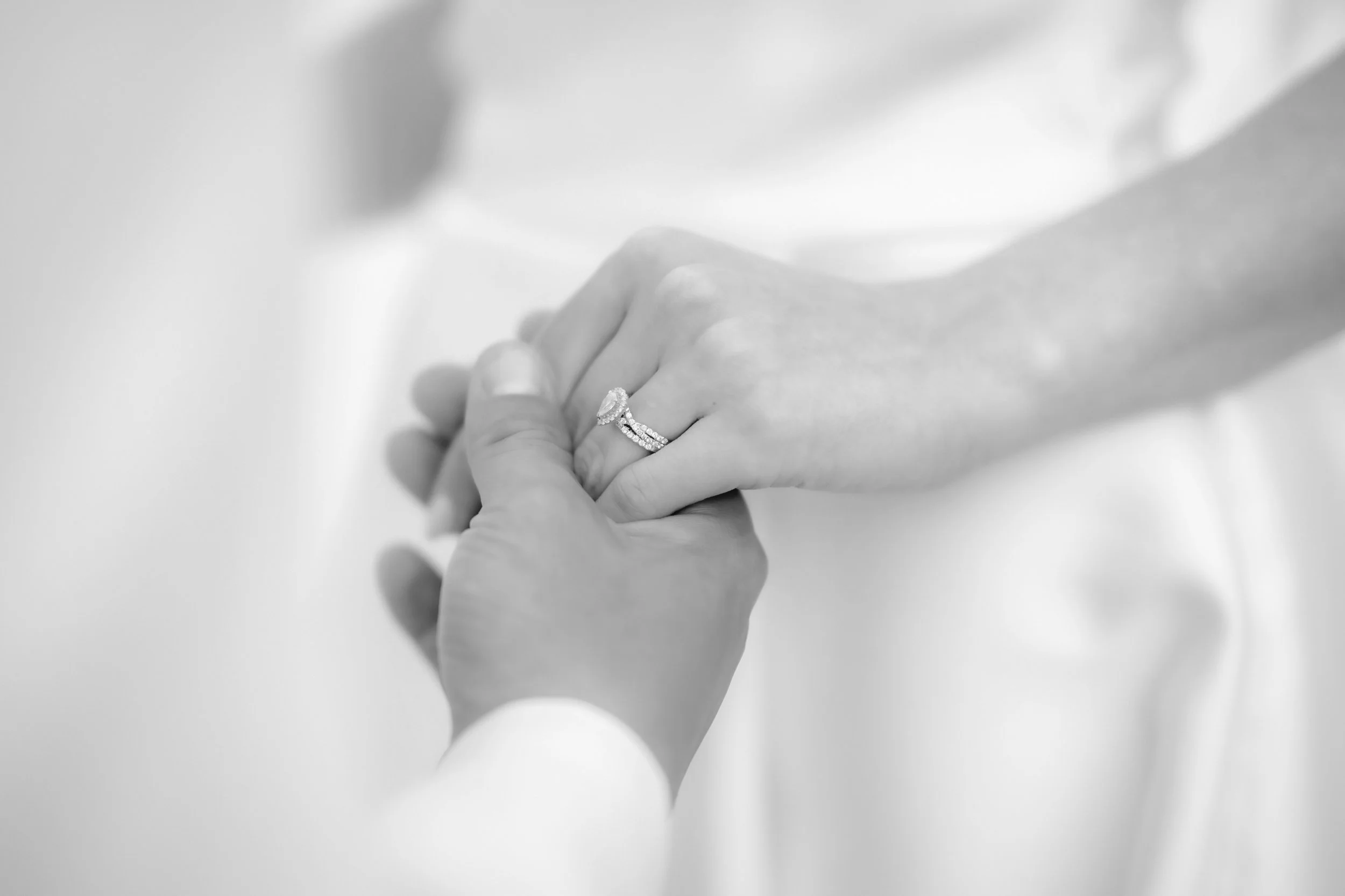 Black and white photograph of a person holding a woman's hand, displaying an engagement ring and wedding band.