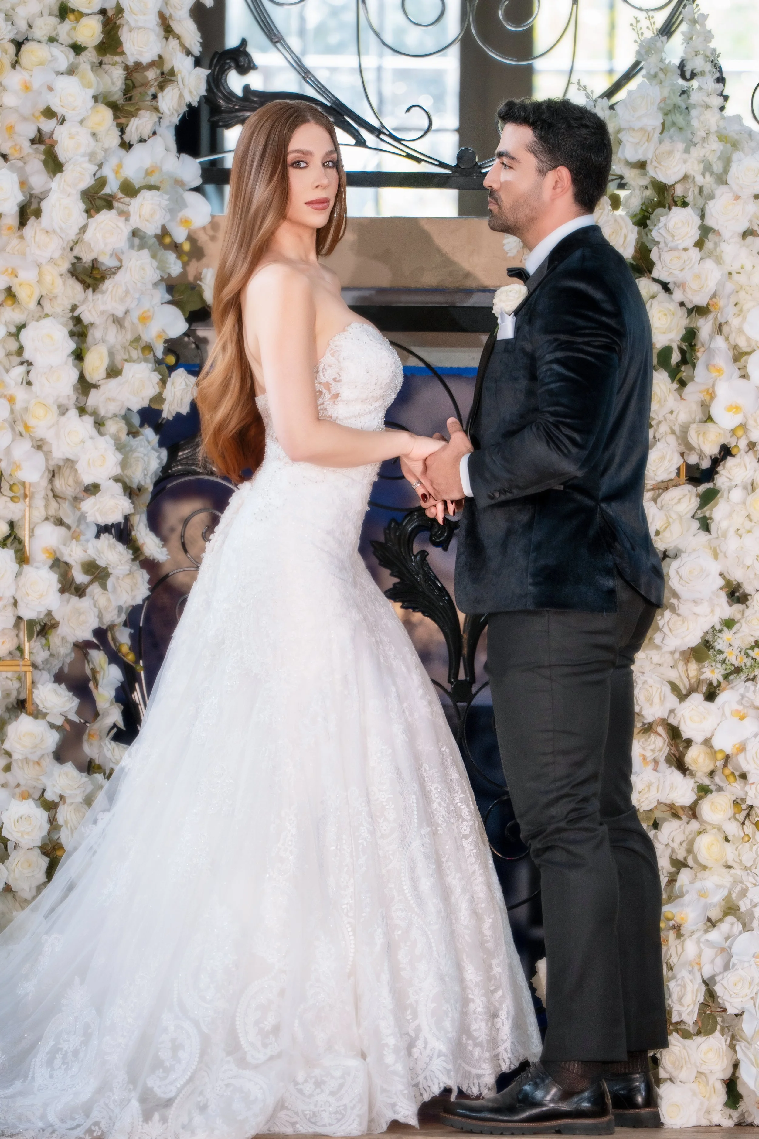 Bride and groom holding hands during their wedding ceremony, standing in front of floral arch with white roses.