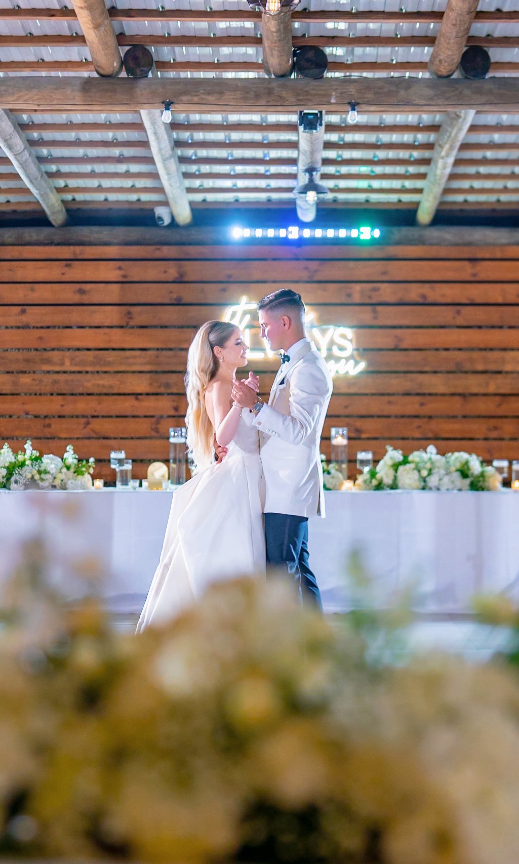 A bride and groom dance together at their wedding reception in a venue with wooden walls and a rustic ceiling. The bride wears a white gown, and the groom wears a white tuxedo jacket with black pants. A neon sign behind them reads 'It’s Always Been Y