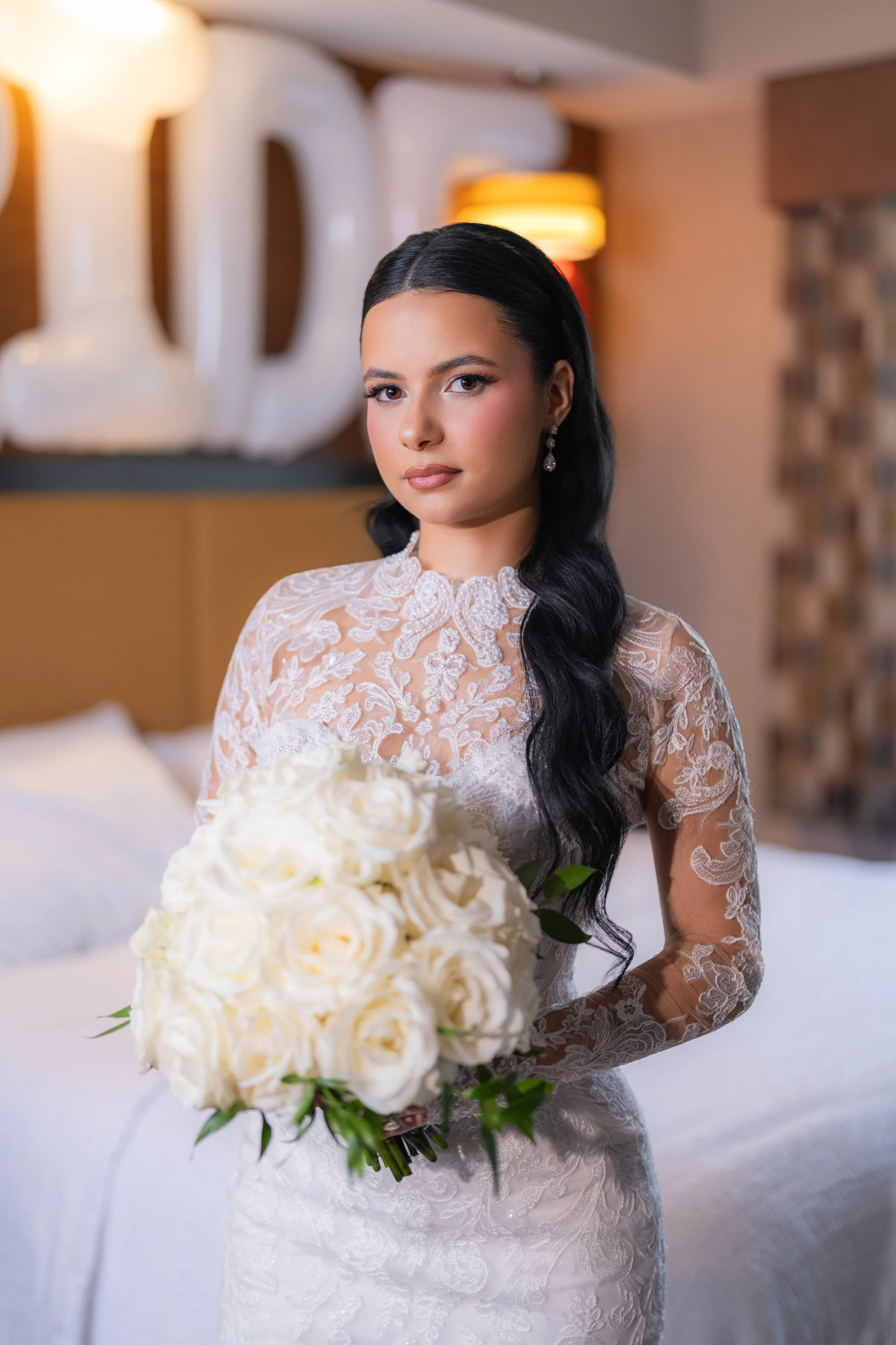 Bride in lace wedding dress holding a bouquet of white roses inside a decorated room.
