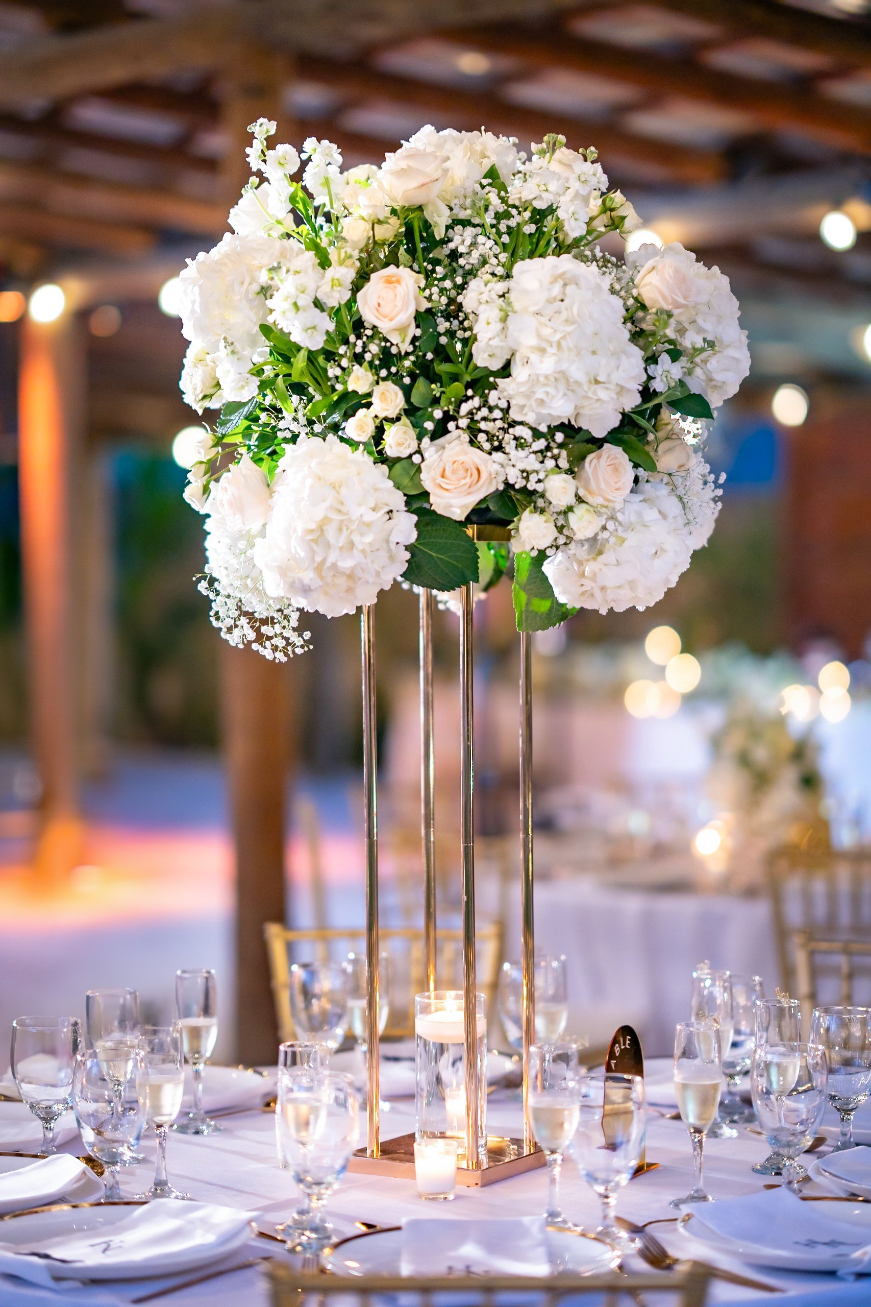 Elegant table setting at a wedding reception with a tall, gold floral centerpiece featuring white roses, hydrangeas, and baby's breath, surrounded by champagne glasses, candles, and gold chairs.