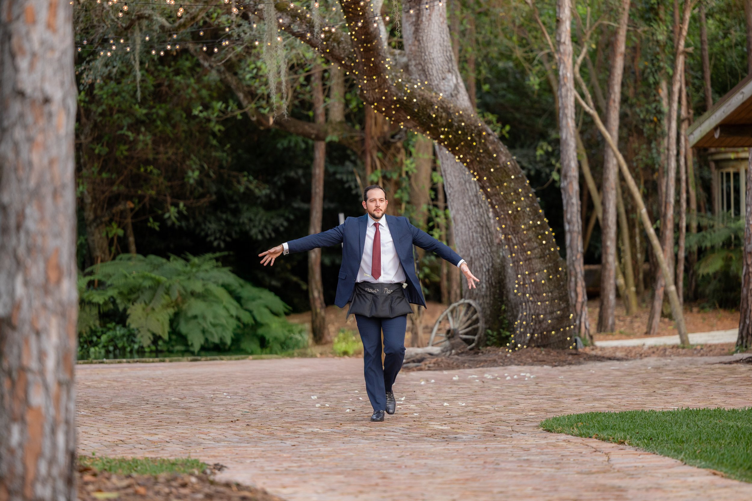 A man in a blue suit with a red tie runs with his arms outstretched on a brick walkway in a wooded outdoor setting, with string lights wrapped around a large tree in the background.